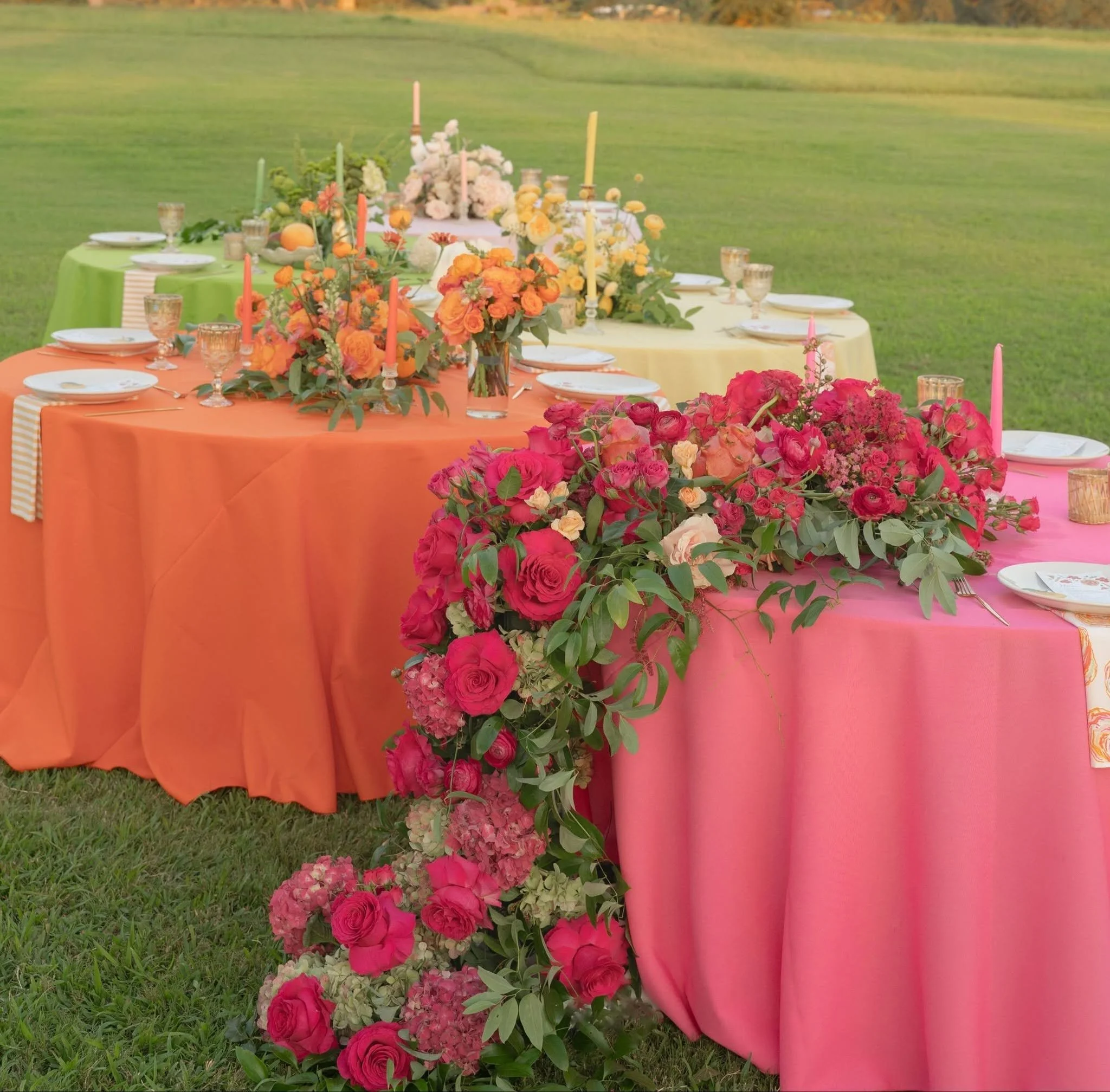 Colorful outdoor table decorated with pink, orange, and white flowers, with candles and glassware, set on grass.