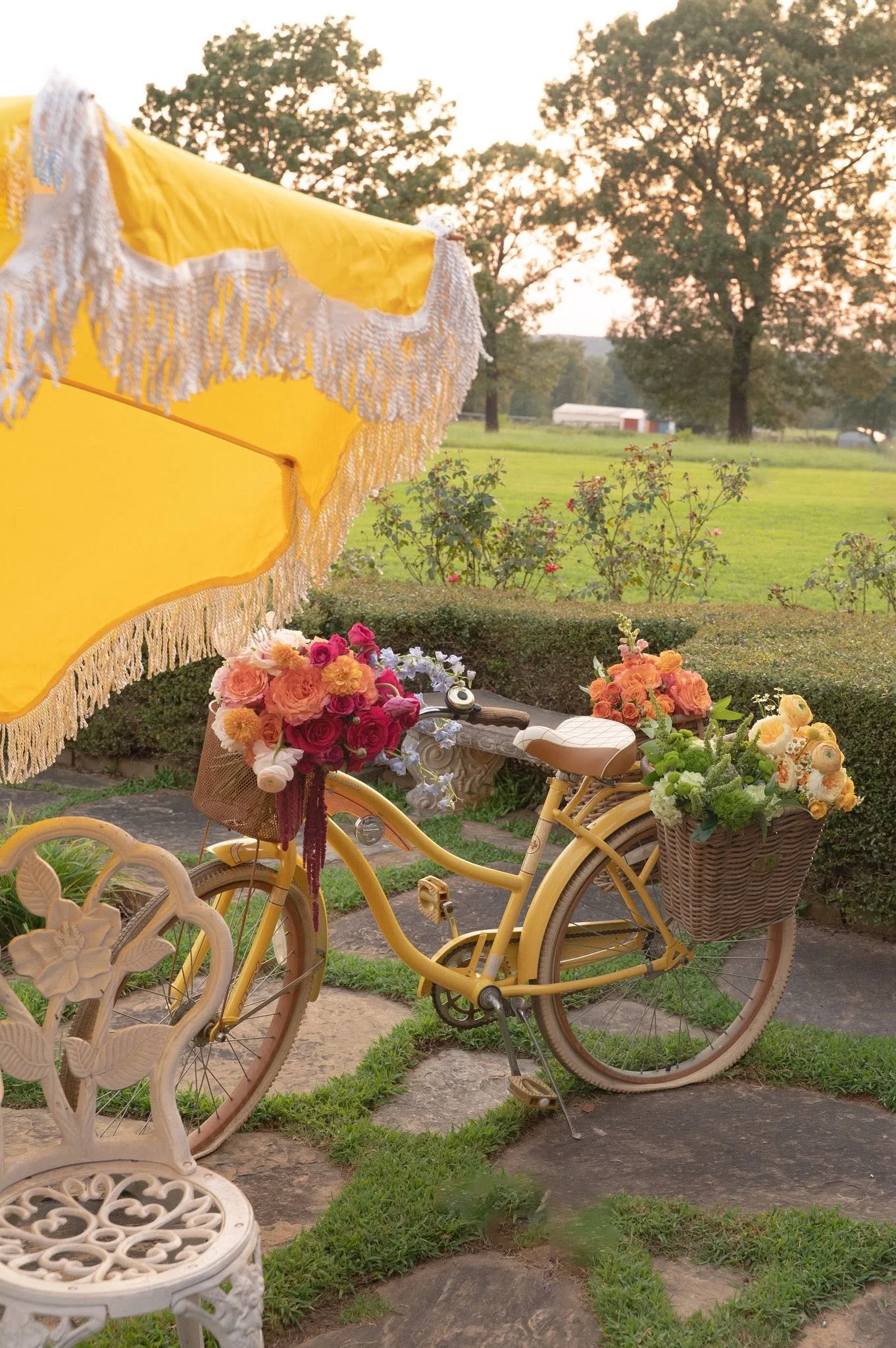 Yellow bicycle with a wicker basket filled with flowers, parked on a pathway with wrought iron garden furniture and a yellow umbrella with white fringe in a garden setting at sunset, with trees and a barn in the background.