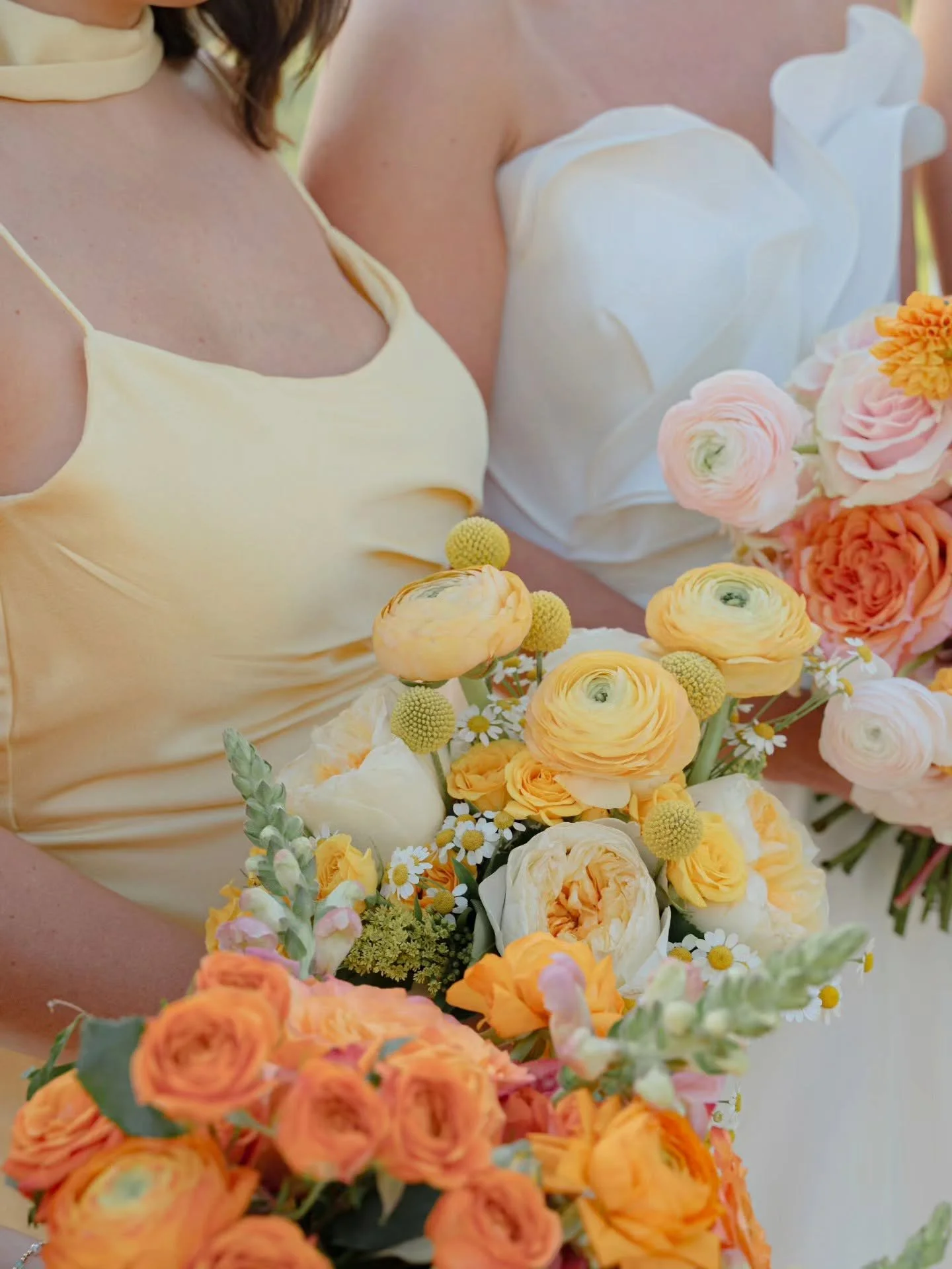 Part of two women in light-colored dresses holding a large bouquet of yellow, orange, and pink flowers with various types of blooms.