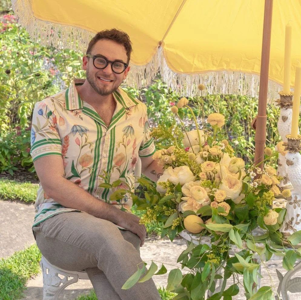 A man with glasses and a patterned short sleeve shirt sitting outdoors at a table with a floral centerpiece, under a yellow umbrella, smiling at the camera.