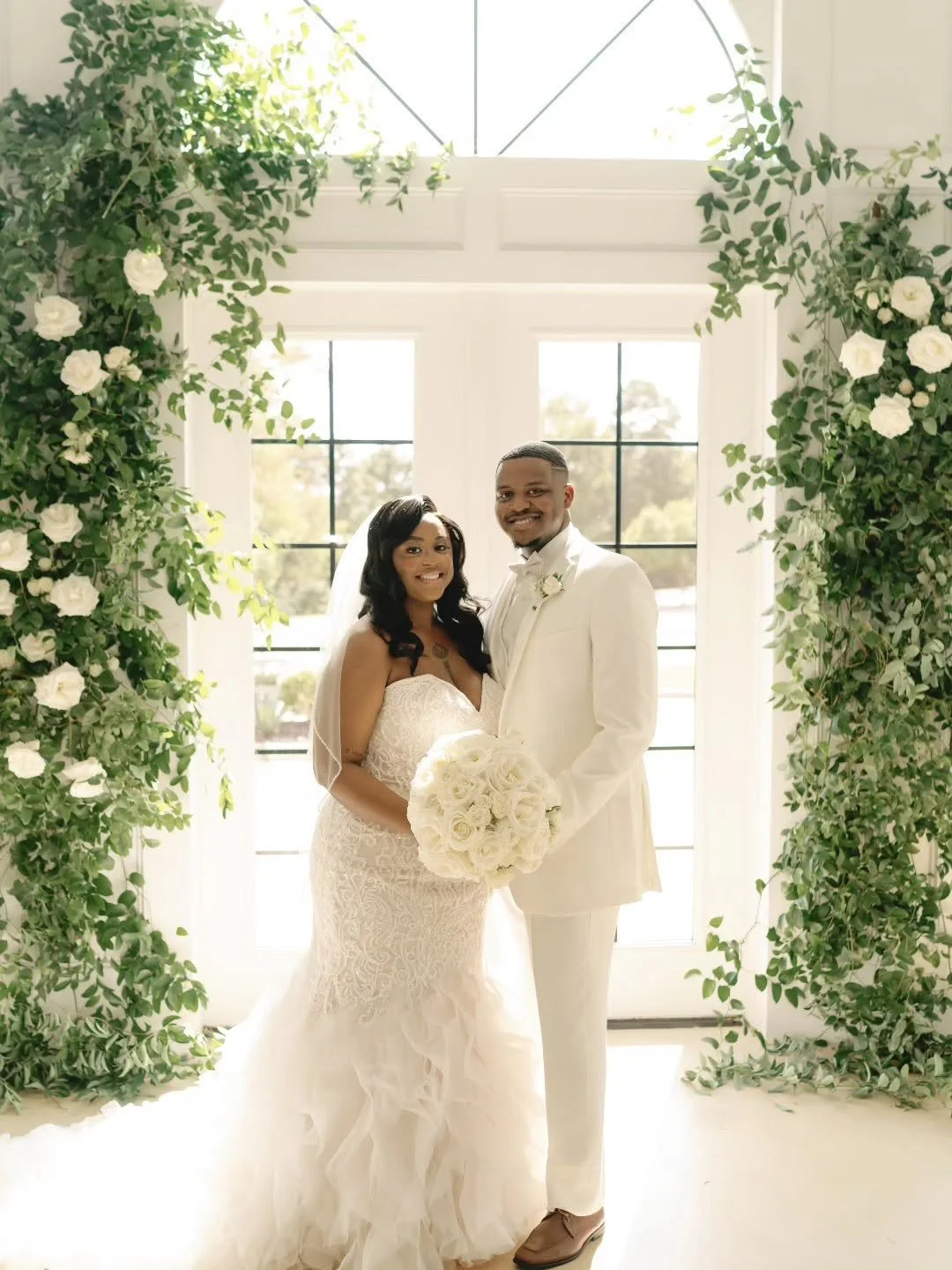 A newlywed couple smiling and holding a bouquet of white roses, standing in front of a large window with floral decorations around the frame, during their wedding celebration.