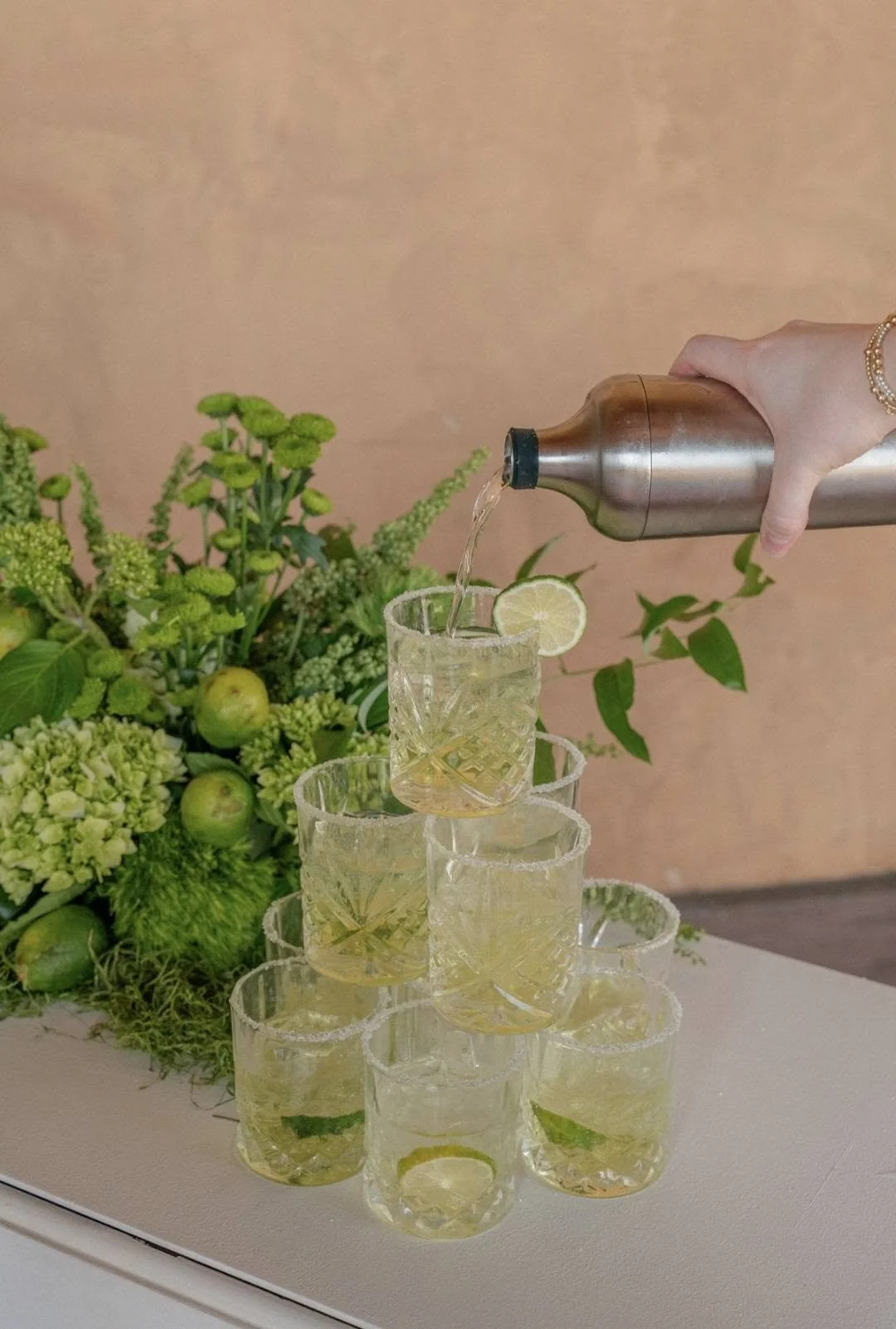 A hand pouring a beverage into a glass with a lemon slice, stacked glasses with lemon slices, a backdrop of green flowers and apples.