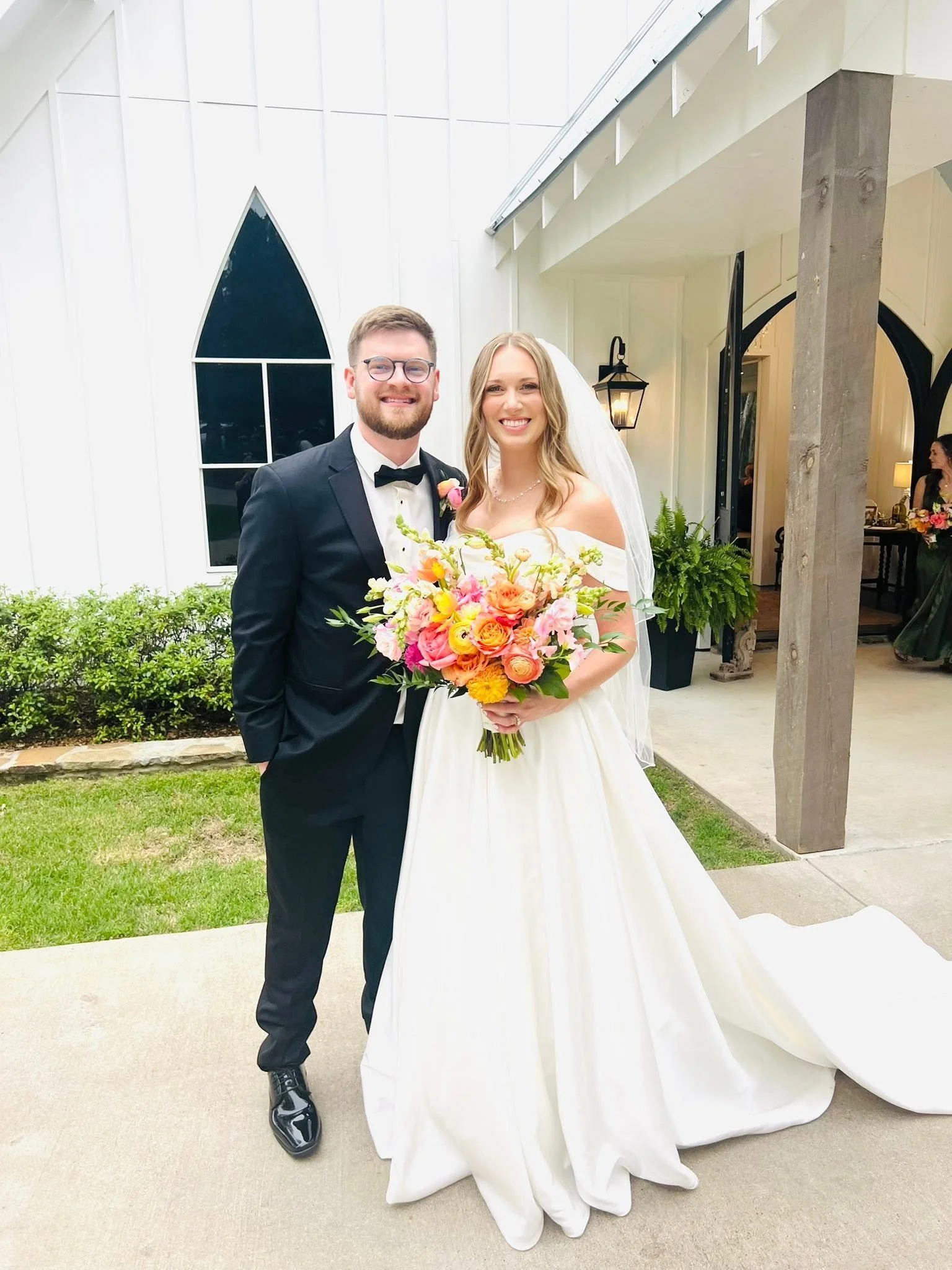A newly married couple standing outside of a church, smiling for a photo. The groom is wearing a black tuxedo, glasses, and a bow tie. The bride is dressed in a white wedding gown, holding a bouquet of colorful flowers.