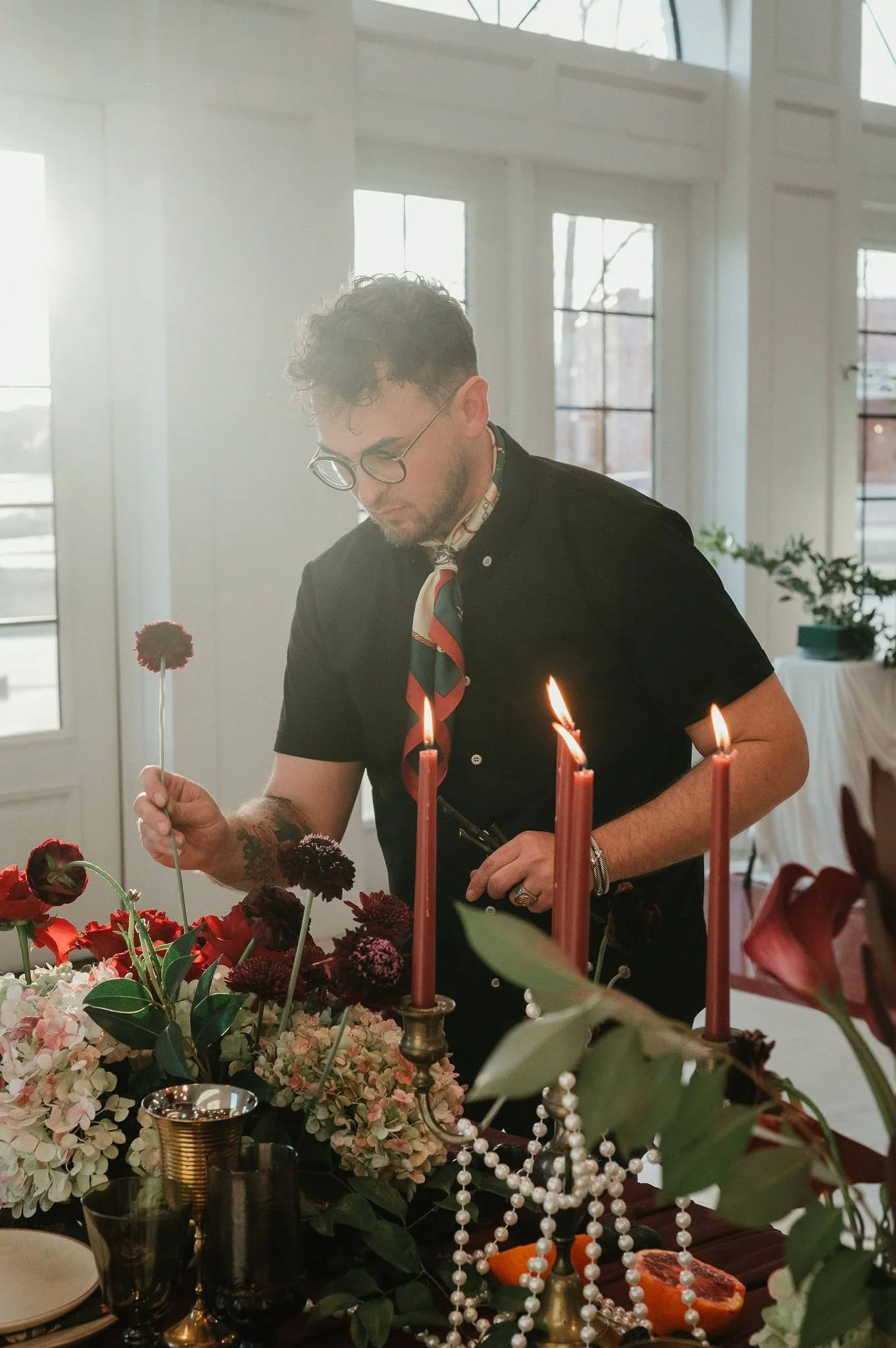 A man with curly hair, glasses, and tattoos, wearing a black shirt and colorful scarf, is lighting a candle on a table decorated with flowers, candles, and jewelry. The room has large windows with sunlight streaming in.