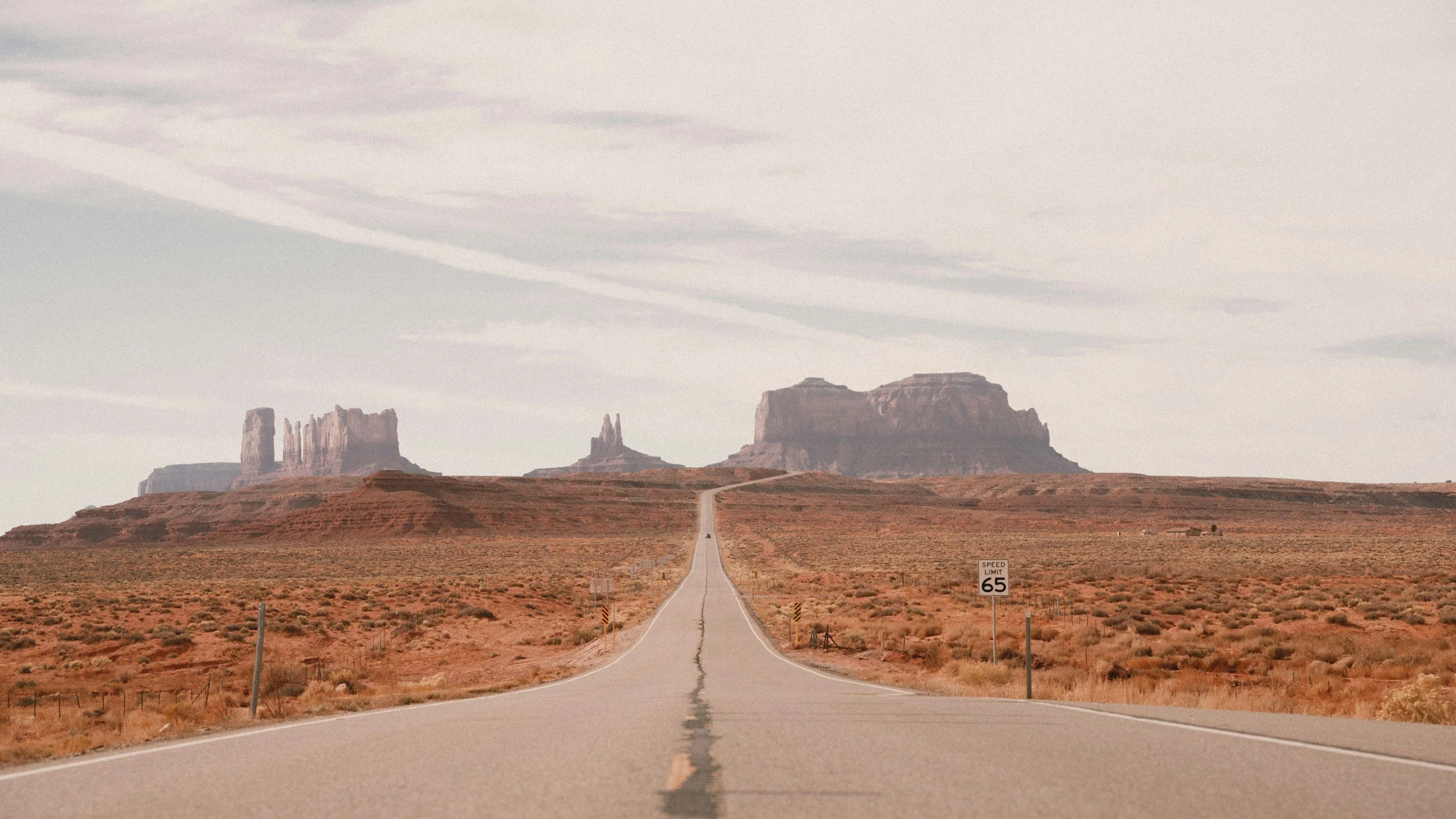 A long straight road leading towards distant rock formations in a desert landscape under a cloudy sky.