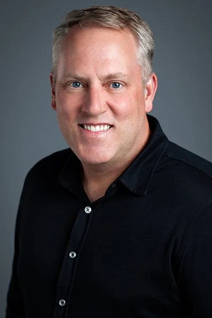 Headshot of a smiling man with short gray hair, wearing a black button-up shirt, against a gray background.