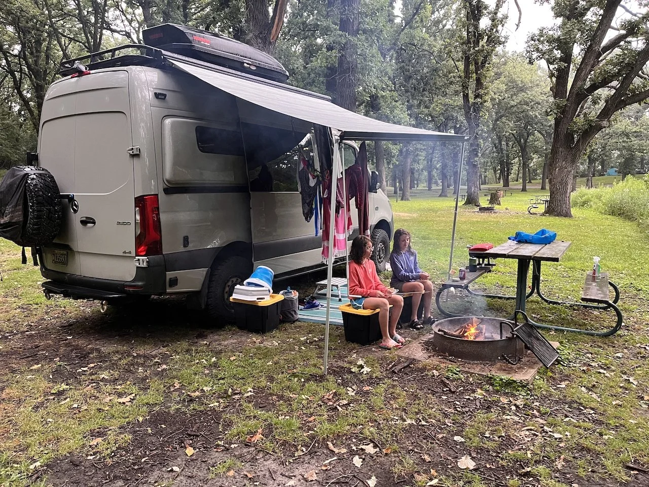 A campsite with a camper van in a wooded park, two children sitting near a campfire, and outdoor furniture and supplies around.