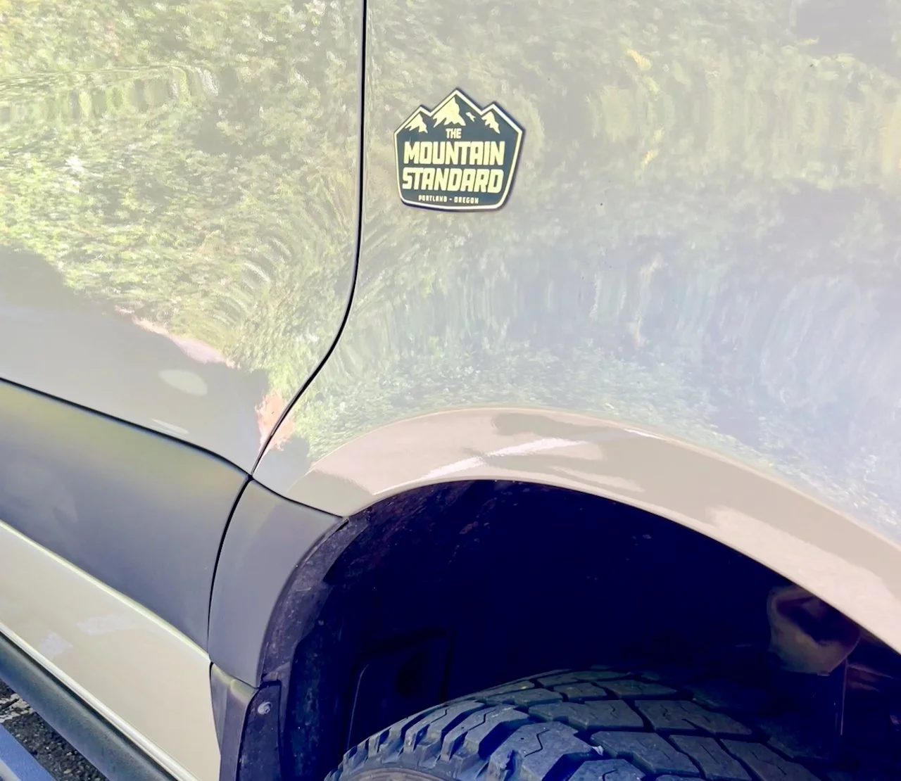 Close-up of a silver vehicle's front side showing a black and white sticker that says "The Mountain Standard" with mountain graphics, located near the front wheel arch.