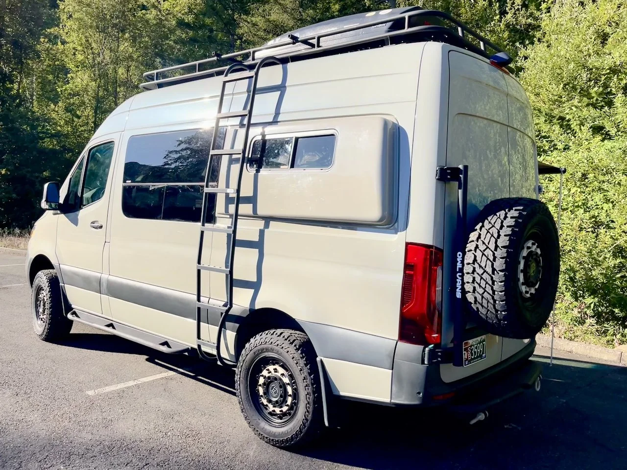 A Sprinter van with a roof rack, ladder, and spare tire parked on a campsite with trees in the background.