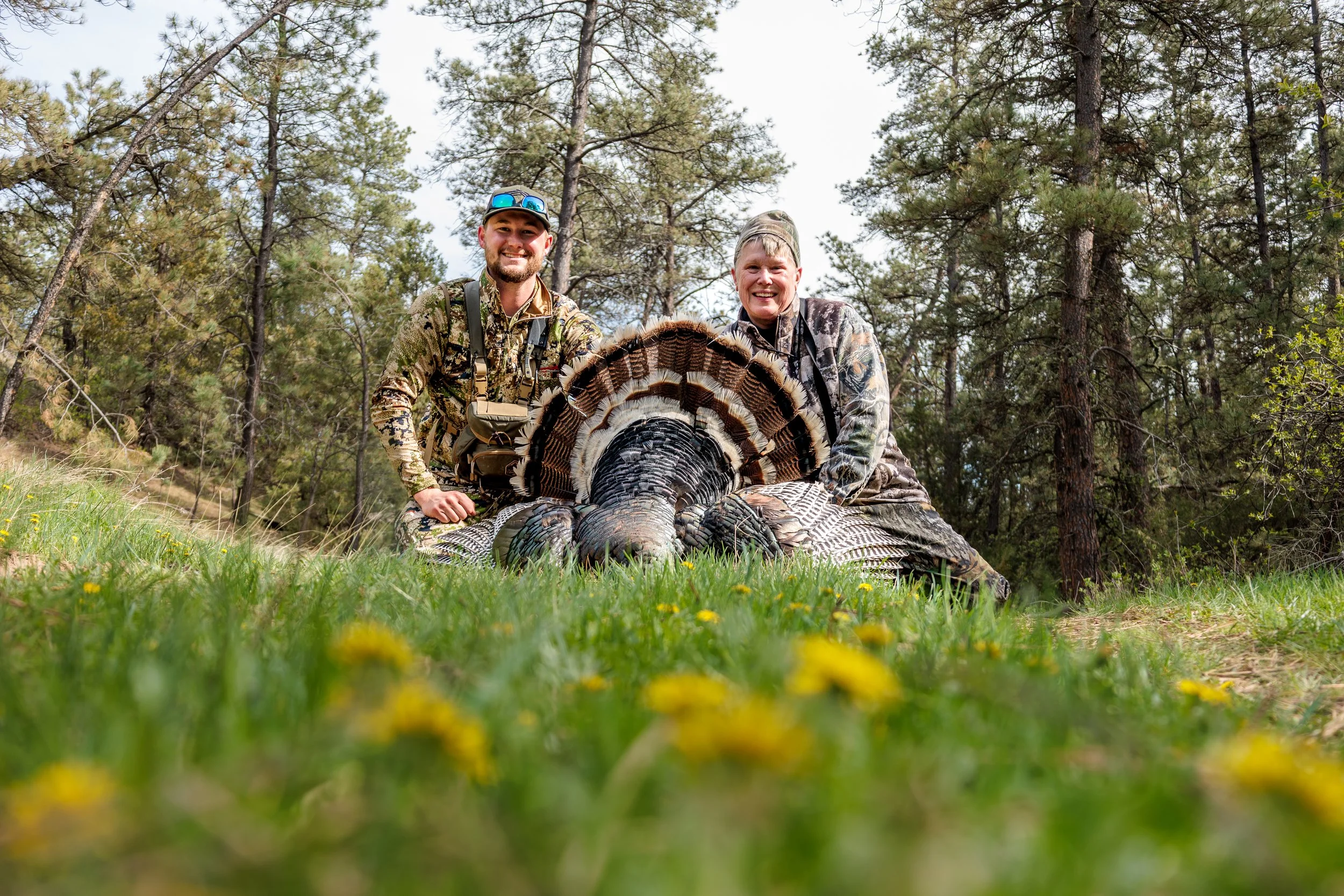 Two men in camouflage clothing smiling next to a dead turkey in a forested area with tall trees and green grass.