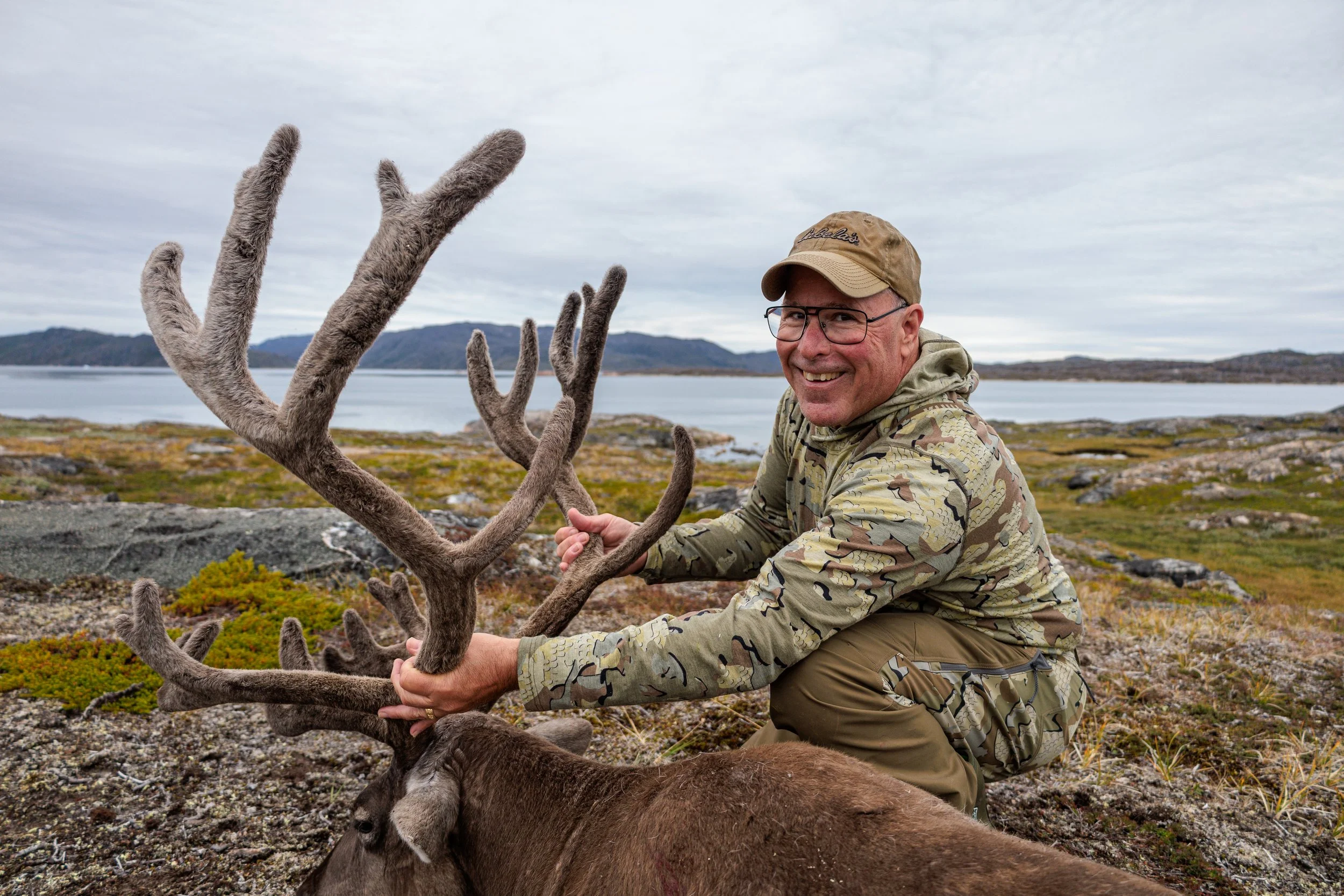 A man wearing camouflage clothing, glasses, and a tan cap is kneeling outdoors, holding the antlers of a reindeer. The landscape features a body of water, rocky terrain, and distant hills or mountains under a cloudy sky.