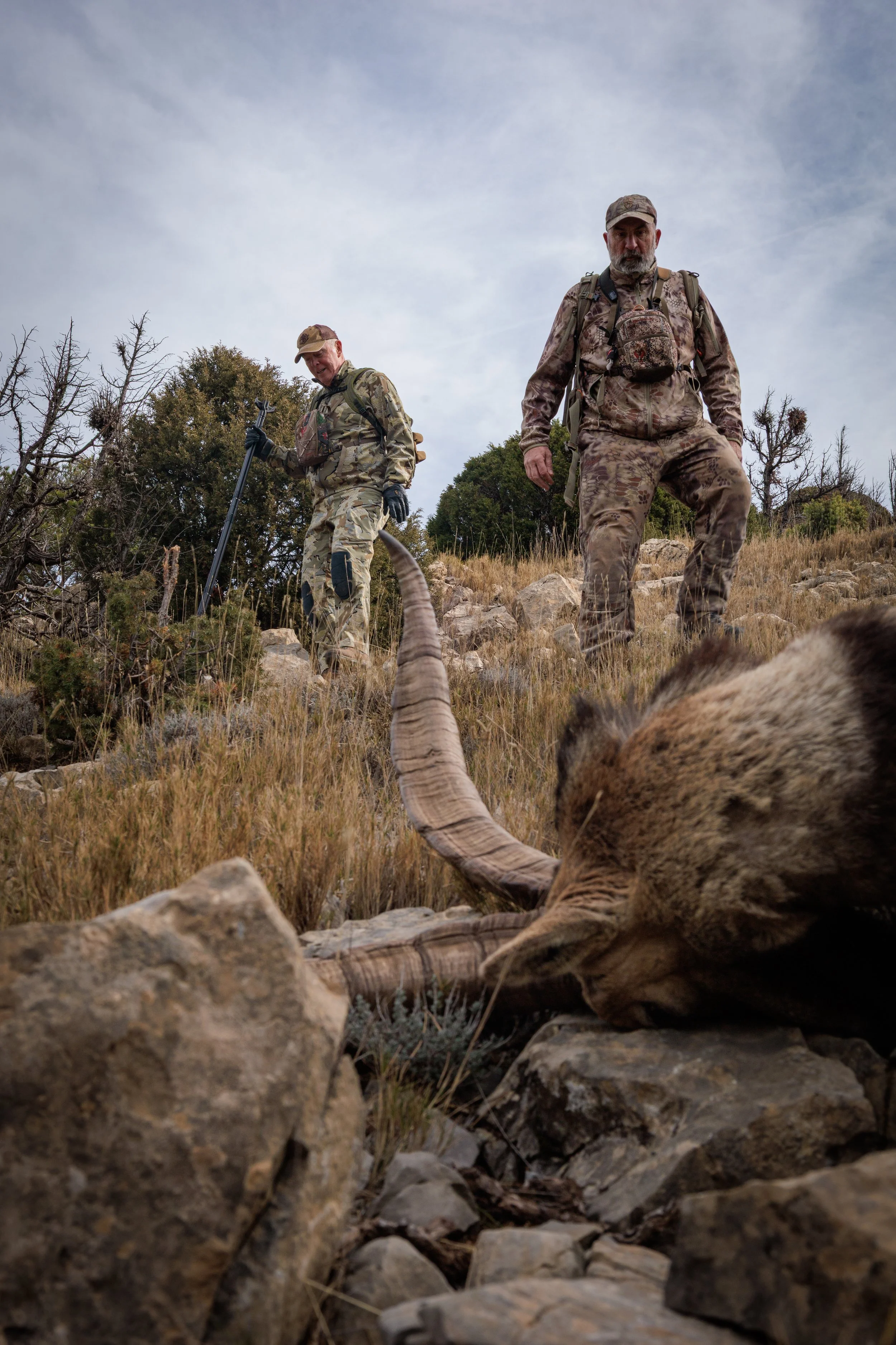 Two men in camouflage clothing and backpacks standing on a rocky hillside with a dead animal lying on its side in the foreground. One man is smiling and holding a walking stick, while the other looks serious, with a cloudy sky overhead.