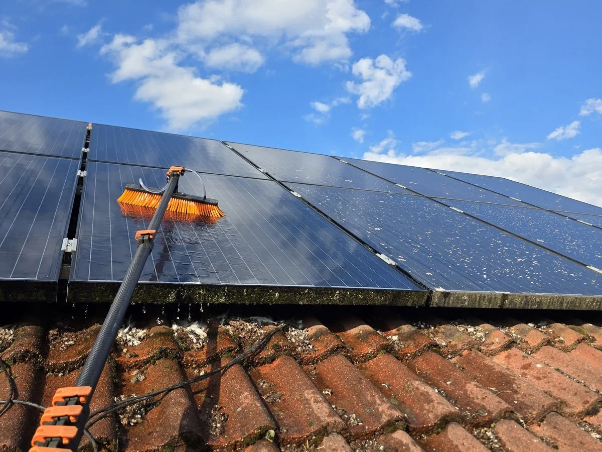 Solar panels on a roof being washed with a brush attached to a long pole under a blue sky with clouds.