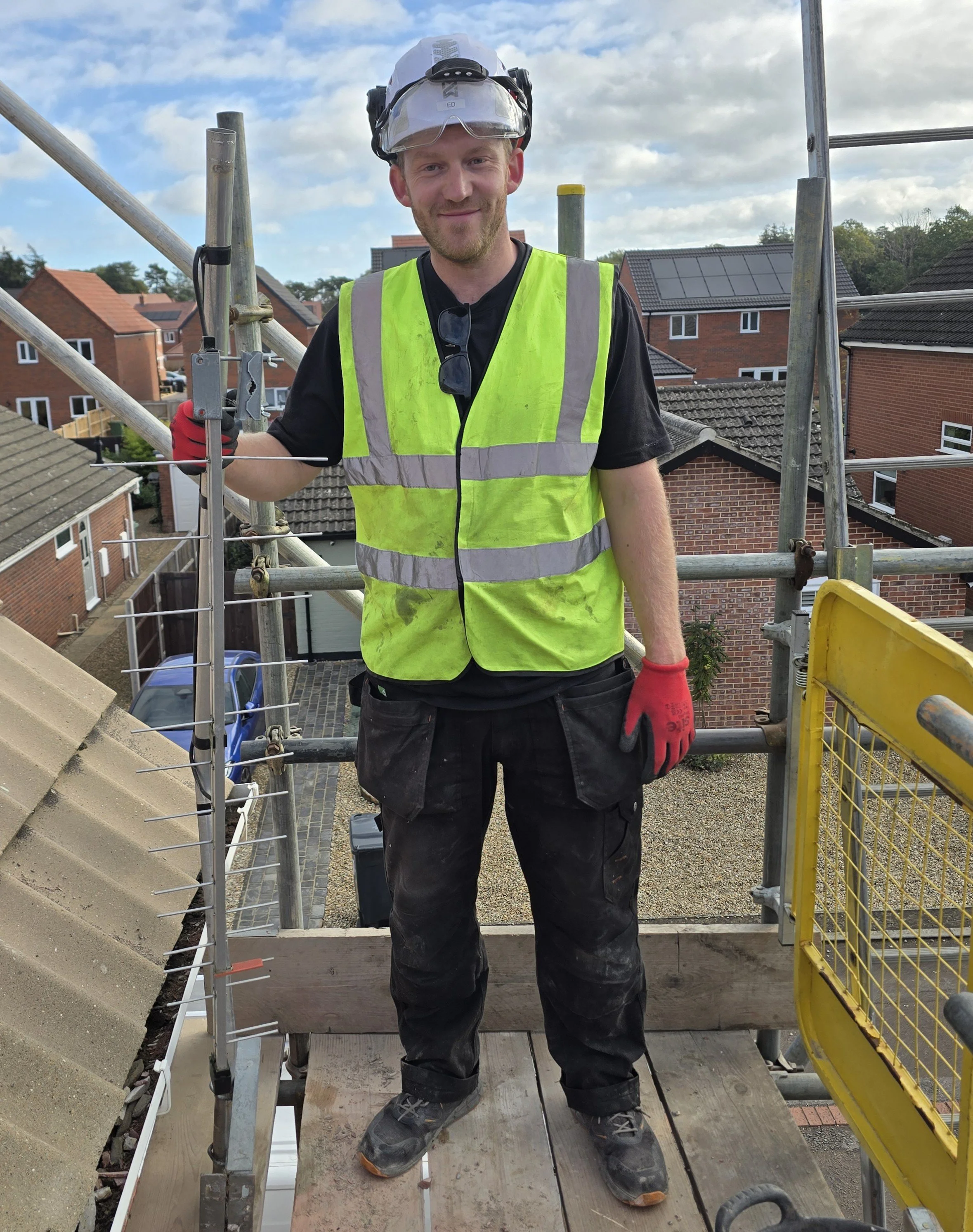 A construction worker standing on a scaffold, wearing a yellow safety vest, helmet with safety glasses, black gloves, and work boots, at a residential building site with houses and a cloudy sky in the background.