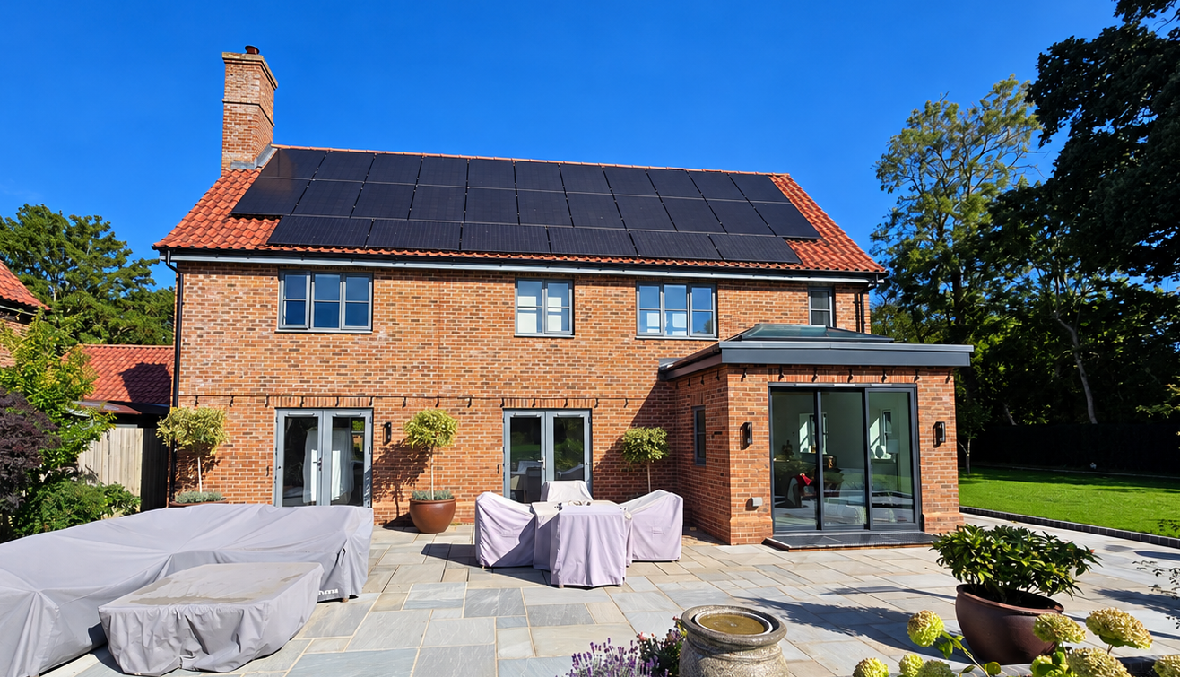 Backyard of a brick house with solar panels on the roof, patio furniture covered with white sheets, potted plants, and trees under a clear blue sky.