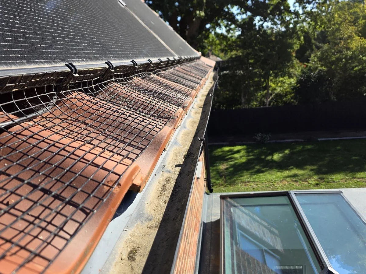Close-up of roof with brick tiles, black mesh guard, and roofing materials, with a garden and trees visible in the background.