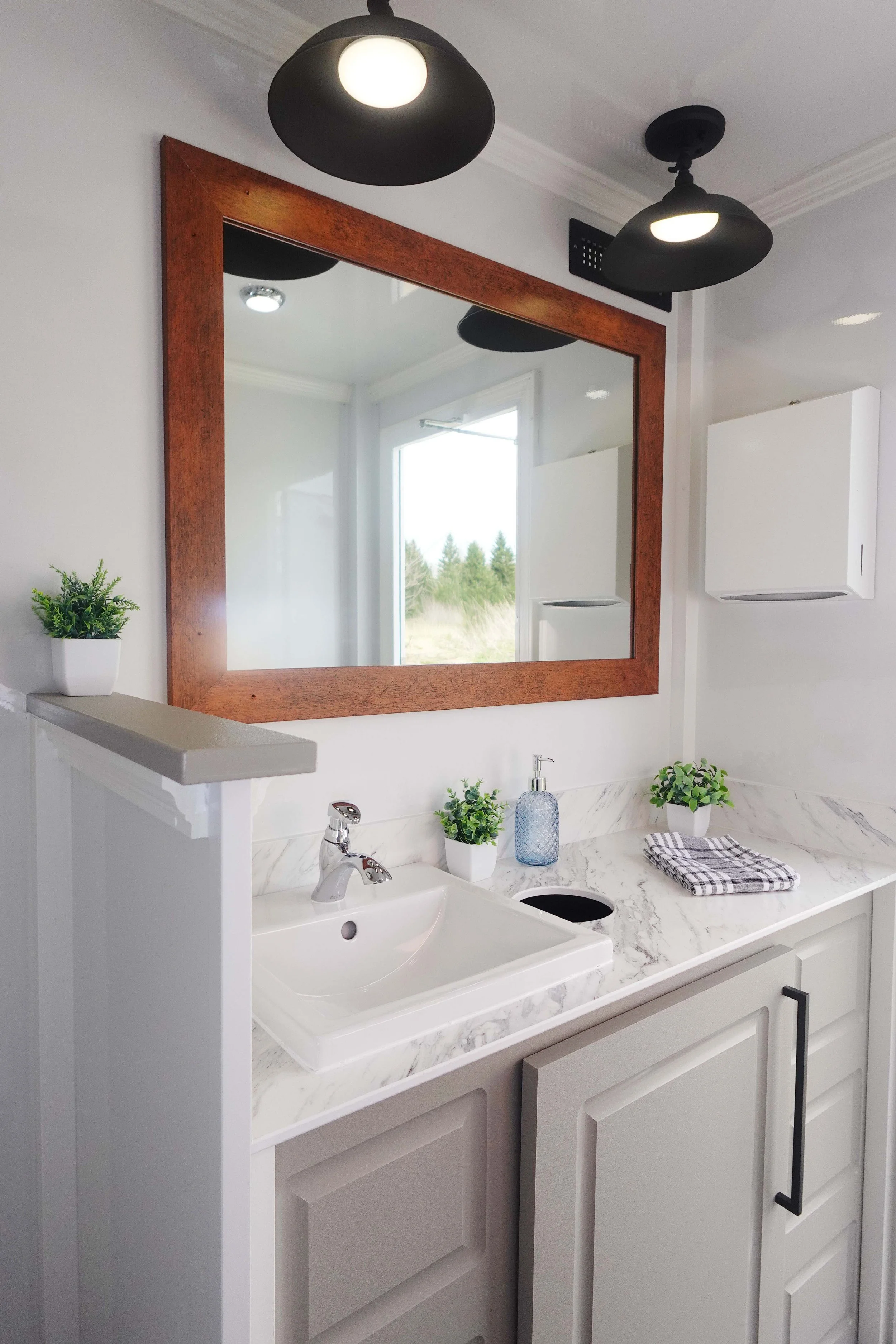 Bathroom vanity with a white marble countertop, a white sink with a silver faucet, and cabinetry with black handles. On the countertop are two small potted plants, a blue glass soap dispenser, and a folded checkered towel. A large mirror with a woode