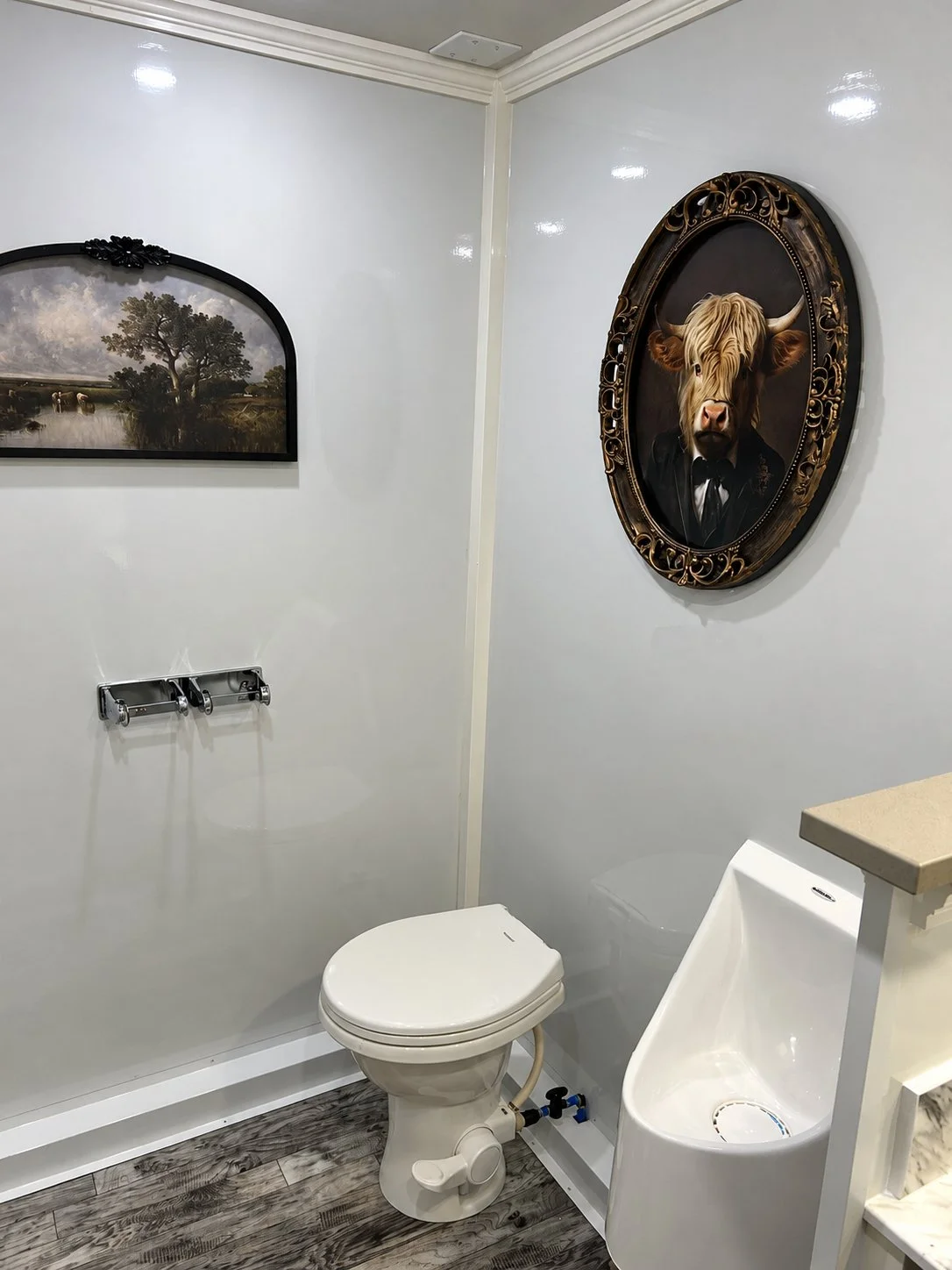 Bathroom with a white toilet, a skid-proof urinal, and two framed pictures on the white wall, one depicting a landscape with trees and water, and the other a portrait of a cow in a suit.