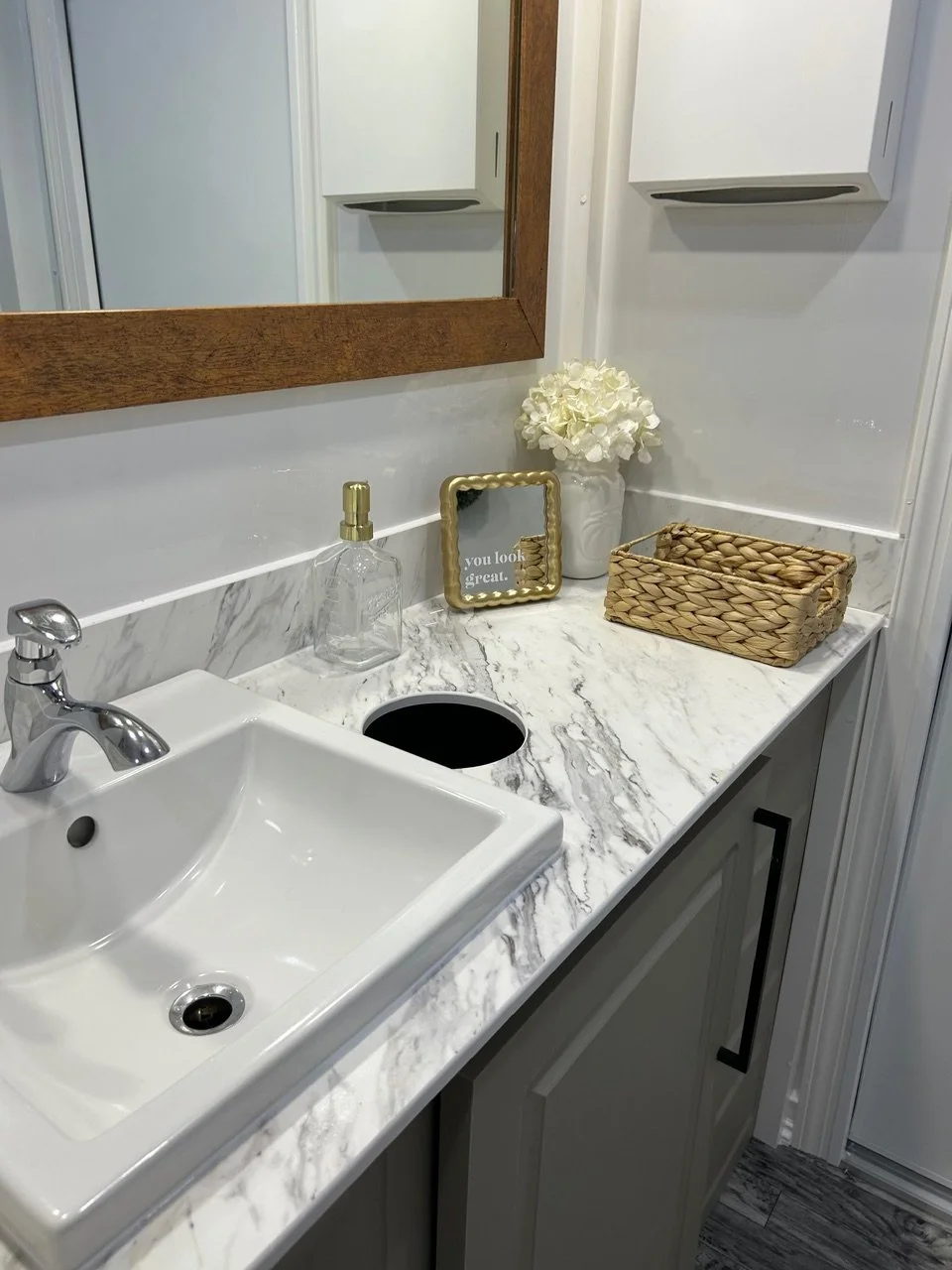 Bathroom vanity with a white sink, marble countertop, a clear soap dispenser with a gold pump, a mirror with a wooden frame, a vase with white flowers, a small framed message that reads "you look great," a woven basket, and a cabinet with black handl