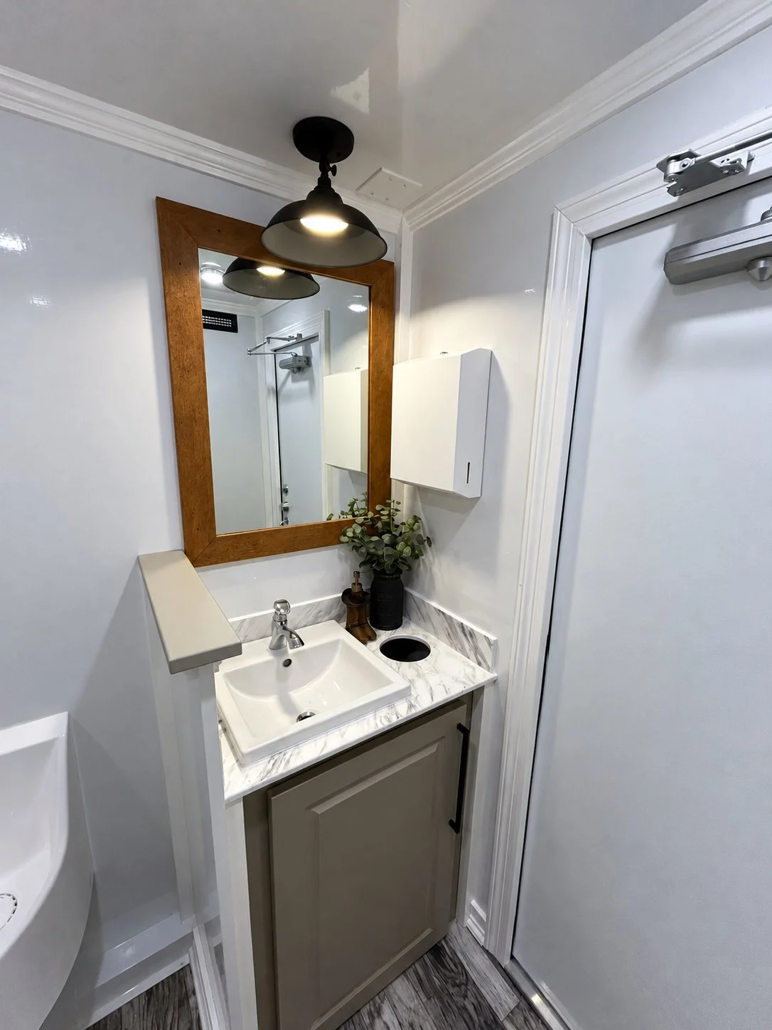 Small bathroom with white walls, a framed mirror, a small sink with a marble countertop, and a black fixture light overhead. There is a white cabinet on the wall and a potted plant next to the sink. A door with a latch is on the right side.