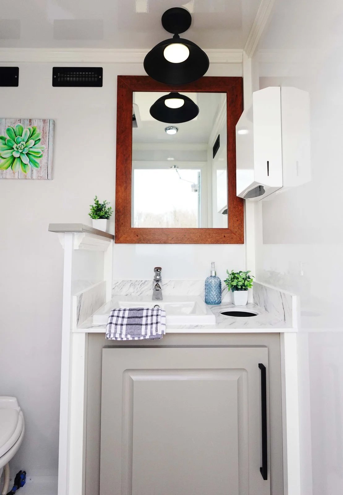 A small bathroom vanity with a marble countertop, a white sink, a mirror with a wooden frame, a black light fixture on the ceiling, and potted plants for decoration.