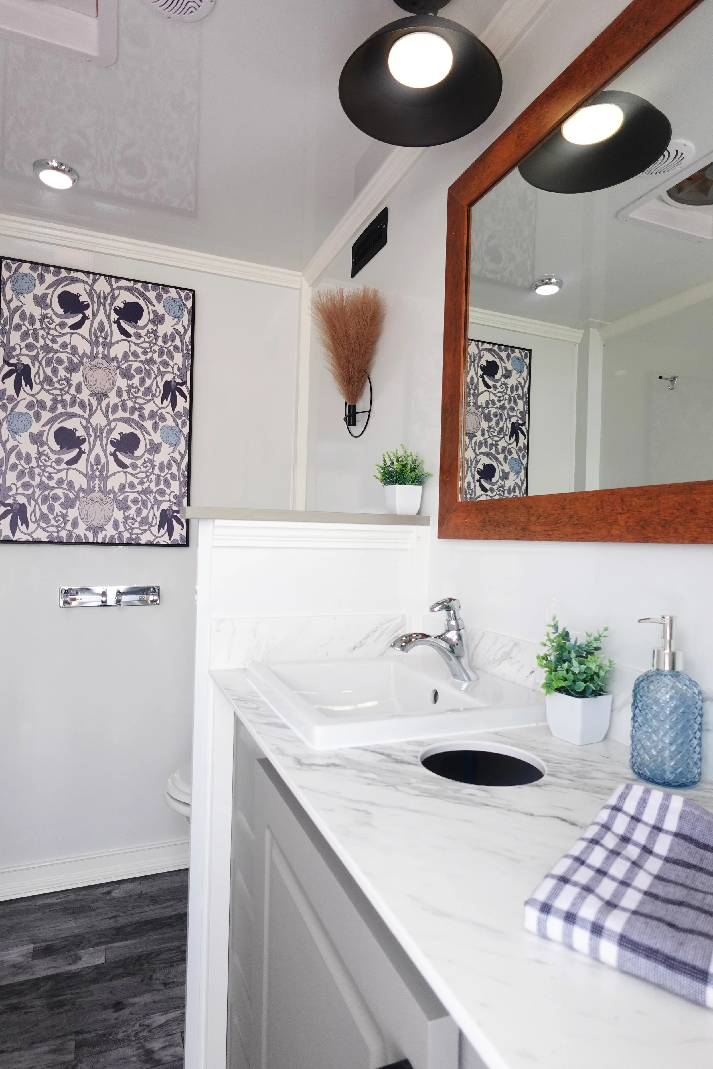 A bathroom with a white sink and marble countertop, a large mirror with a wooden frame, and decorative elements including a small potted plant, a blue vase, a wall-mounted light fixture, and framed artwork with floral patterns, and a checkered towel.