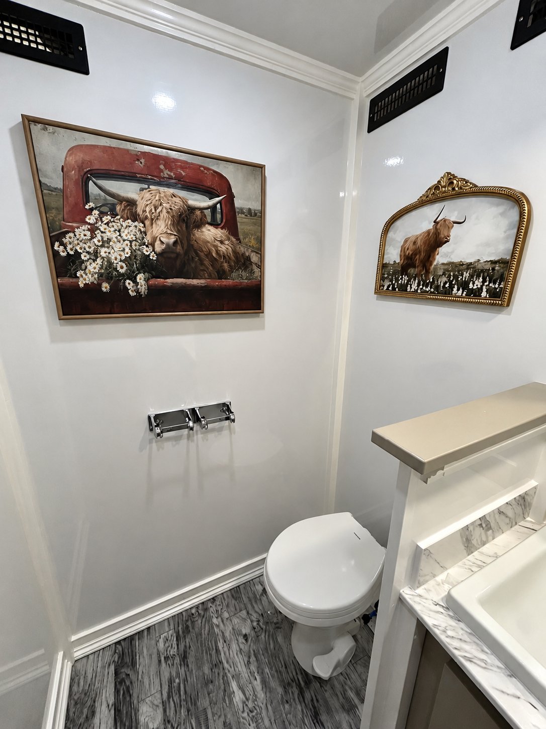 Rustic luxury restroom trailer interior featuring Highland cow artwork, a private toilet, marble-look countertop, and wood-look flooring