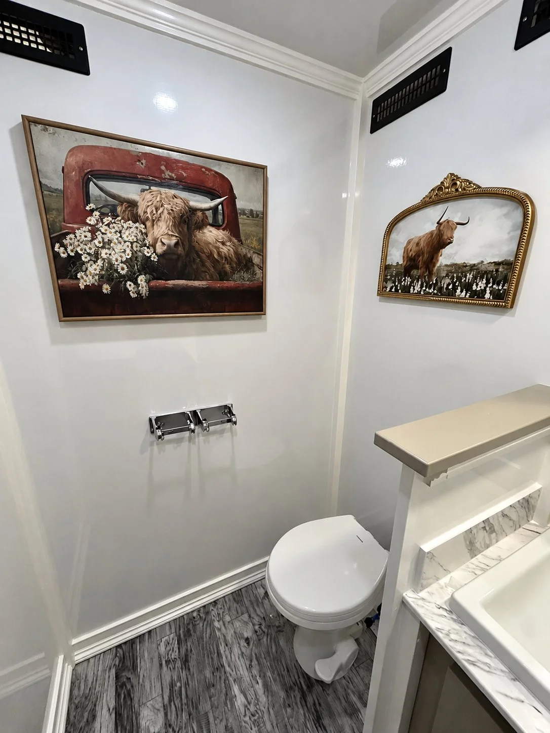 A small bathroom with white walls and a dark wood floor. There are two paintings of highland cattle on the walls, one framed in wood and the other with a gold ornate frame. A toilet is visible, and there is a marble countertop with a sink on the righ