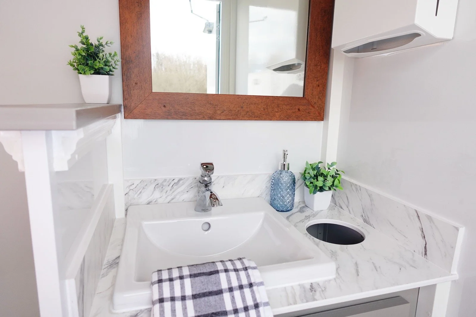 Small bathroom sink with a striped hand towel, a soap dispenser, a potted plant, a mirror, and a window in the background.