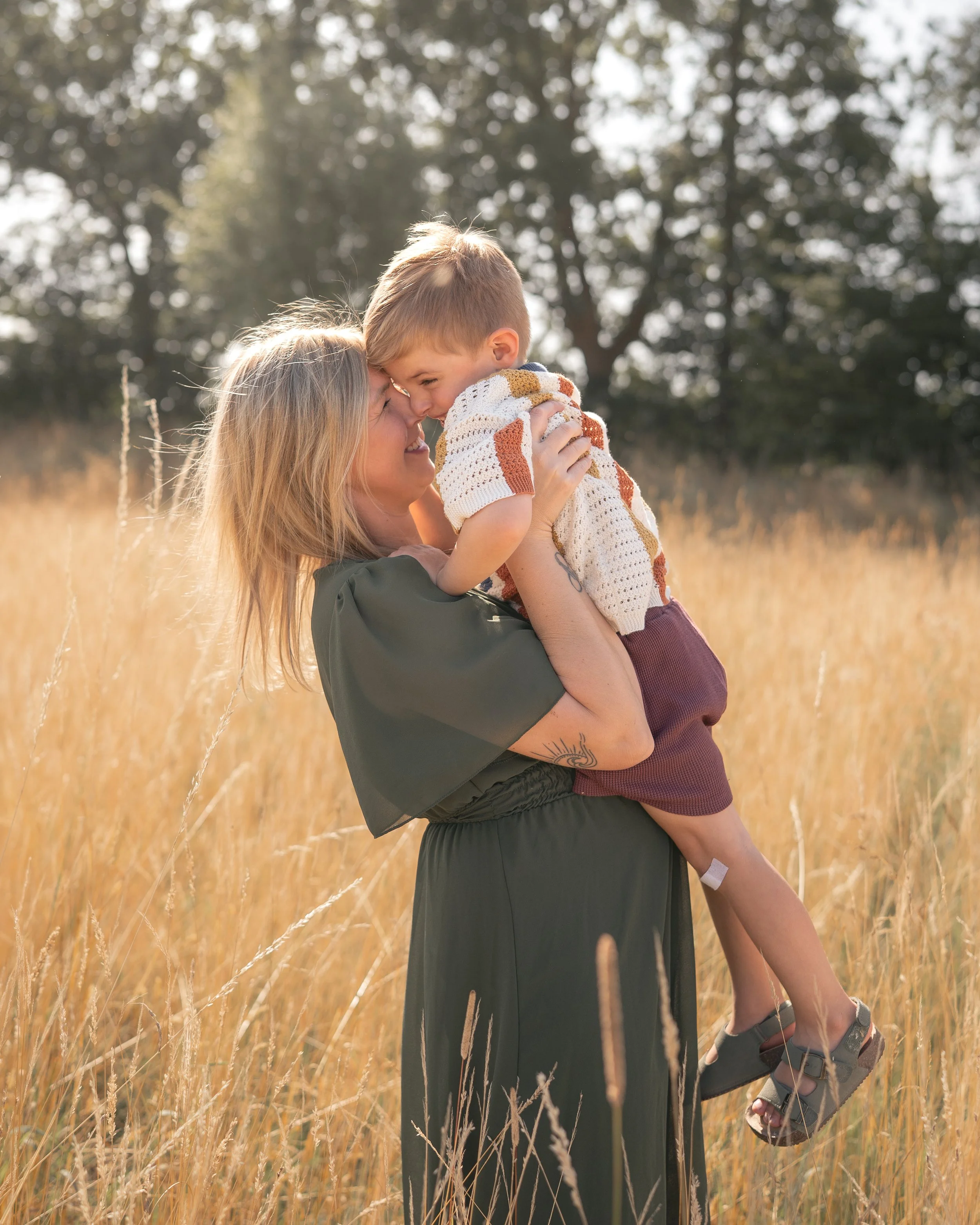 Eine Frau hebt ein lachendes Kind in die Luft in einem goldenen Feld bei Sonnenuntergang, bunte Bäume im Hintergrund.