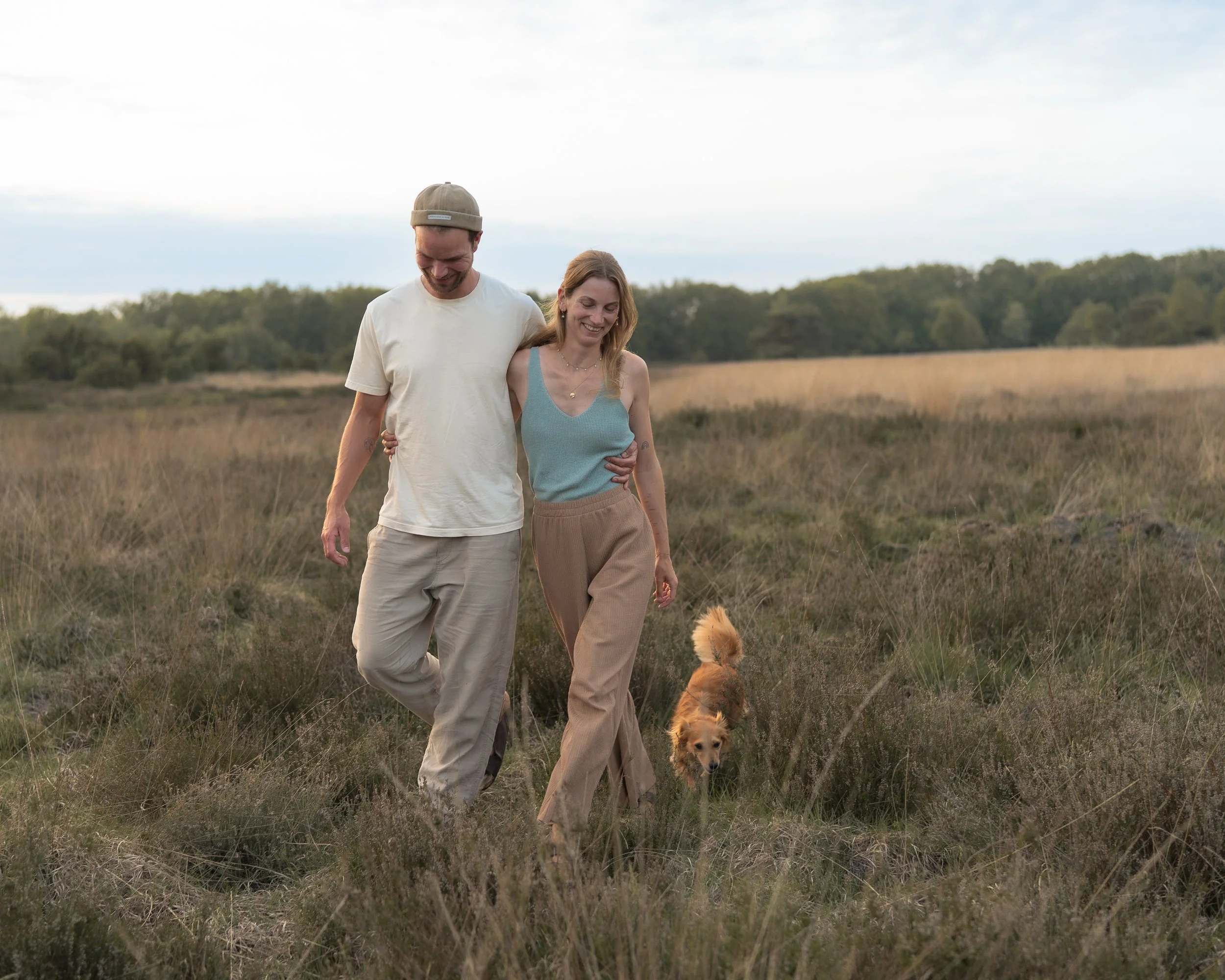 Ein Mann und eine Frau gehen einen Pfad in einer offenen Naturlandschaft entlang, begleitet von zwei Welpen, bei Tageslicht im Freien.
