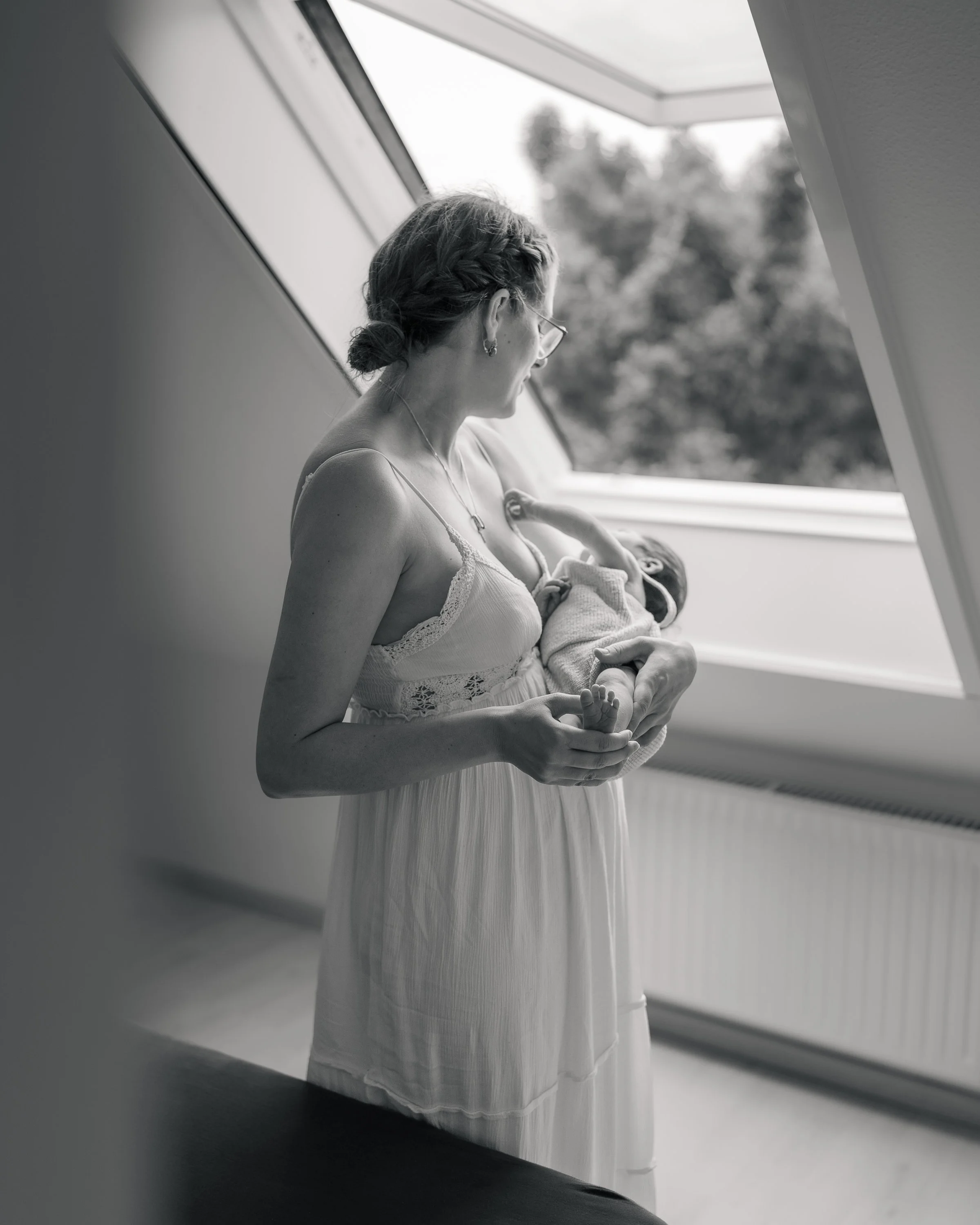 Eine Frau mit Locken im Haar und Brille hält ein Baby im Arm, während sie in einem Raum mit einem großen Fenster steht. Die Frau trägt ein weißes Kleid. Das Bild ist schwarz-weiß.
