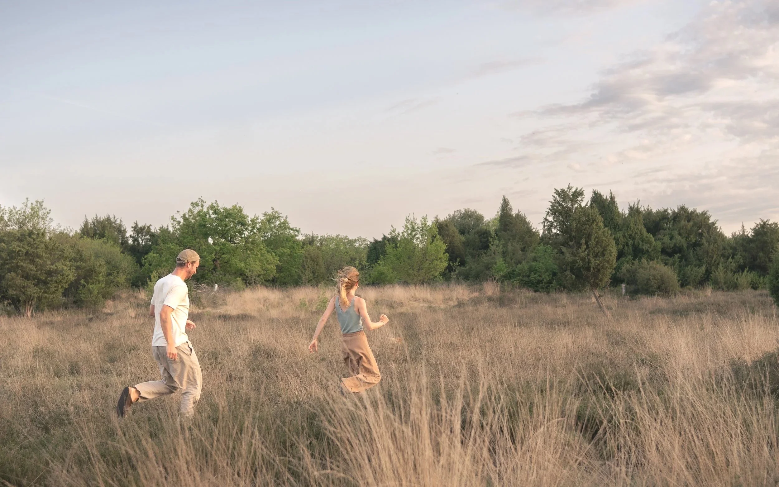 Drei Personen - ein Mann, eine Frau und ein Mädchen - laufen durch ein hohes Grasfeld im Freien bei Sonnenuntergang, mit Bäumen im Hintergrund und einem Himmel mit Wolken.
