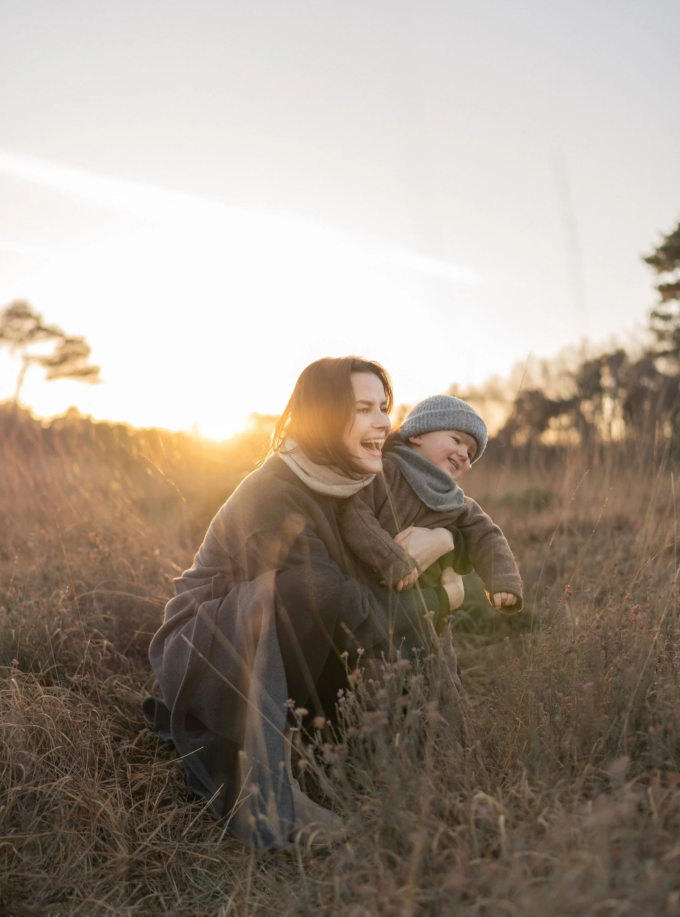 Eine lachende Frau und ein lachender Junge sitzen im Feld bei Sonnenuntergang, umgeben von trockenem Gras und Bäumen im Hintergrund.