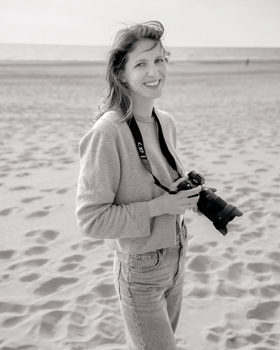 Eine Frau am Strand, hält eine Kamera und lächelt, im Schwarz-Weiß-Bild.