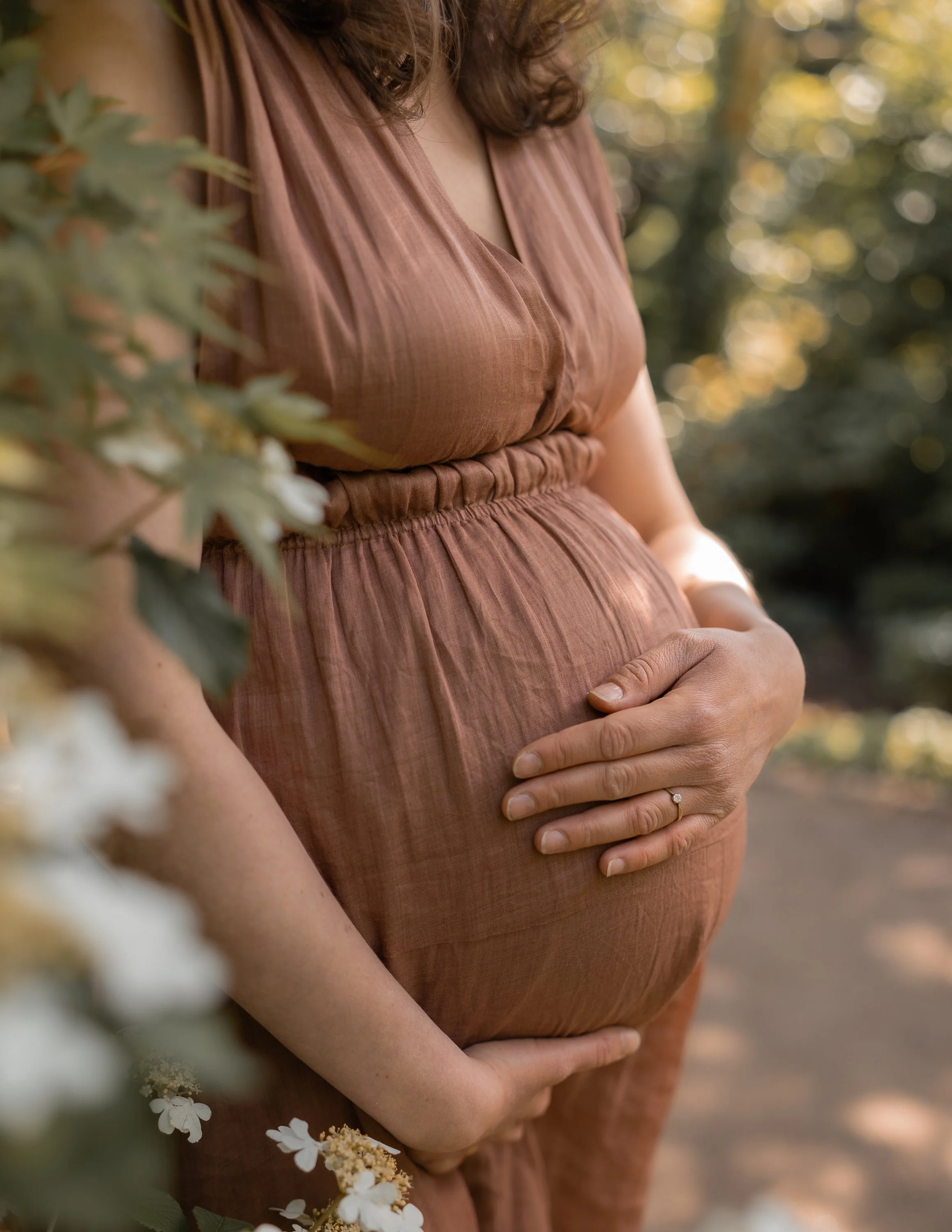 Eine schwangere Frau trägt ein braunes Kleid und hält ihre Hände auf den Bauch, während sie im Freien im Sonnenlicht steht.