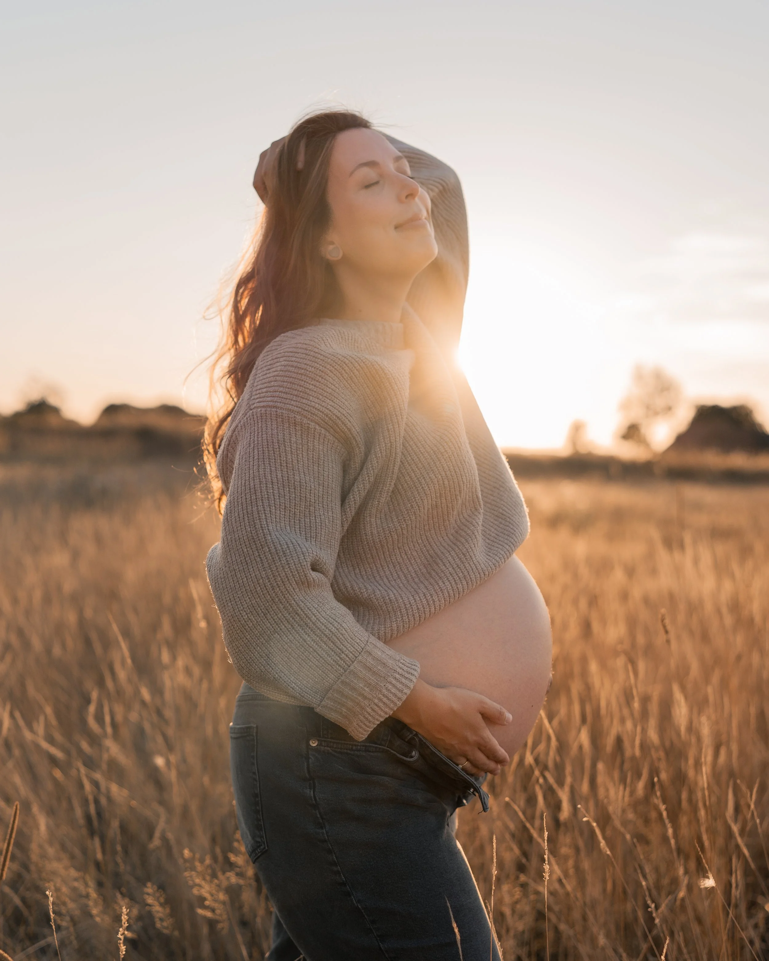 Eine schwangere Frau steht in einem goldenen Feld bei Sonnenuntergang, die Augen geschlossen und ein zufriedenes Lächeln im Gesicht, während sie ihre Hand auf den Bauch legt.
