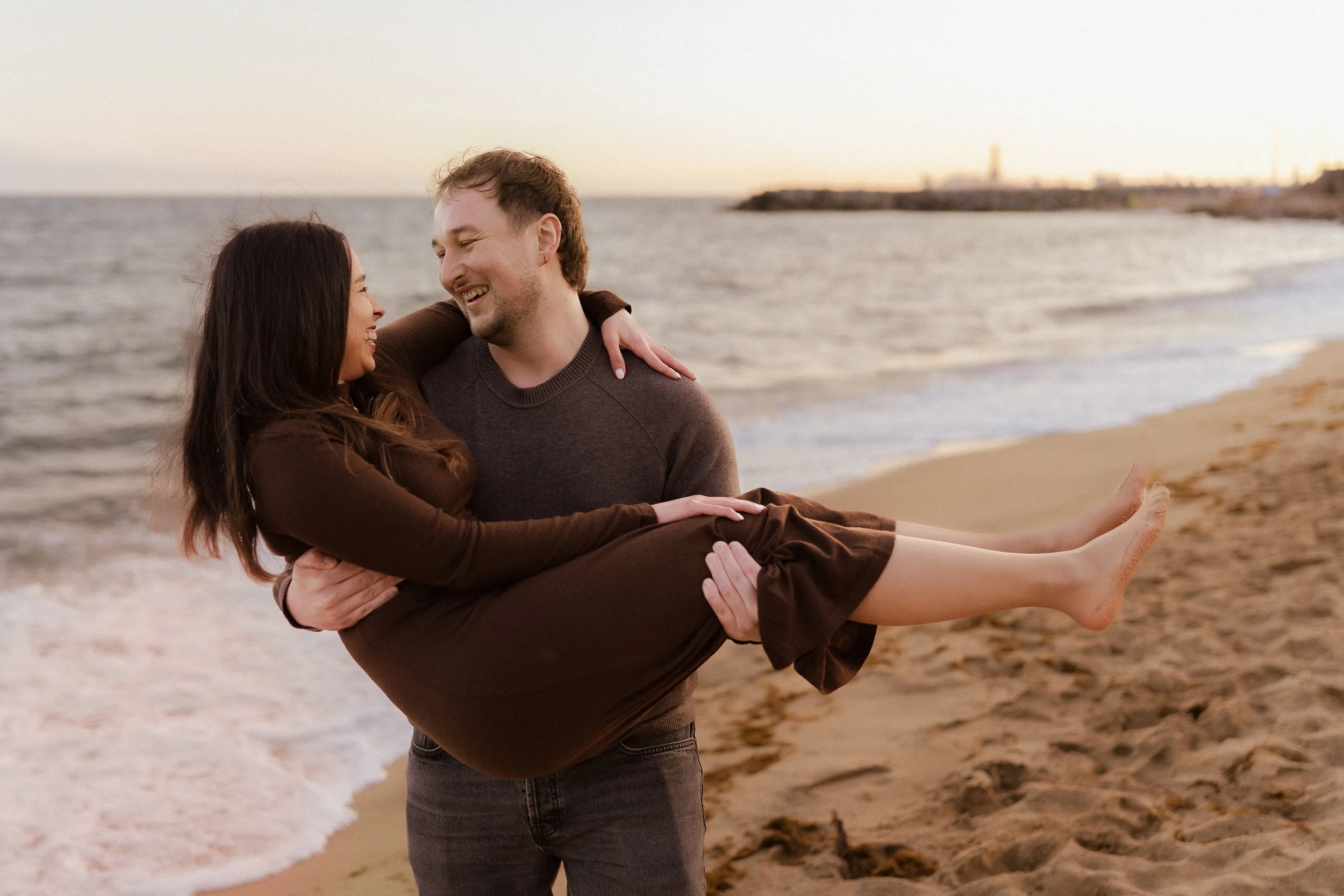 Ein Paar am Strand, der Mann trägt eine graue Sweatshirt, er hält die Frau in den Armen, während sie gemeinsam am Ufer sitzen, im Hintergrund der Ozean bei Sonnenuntergang.