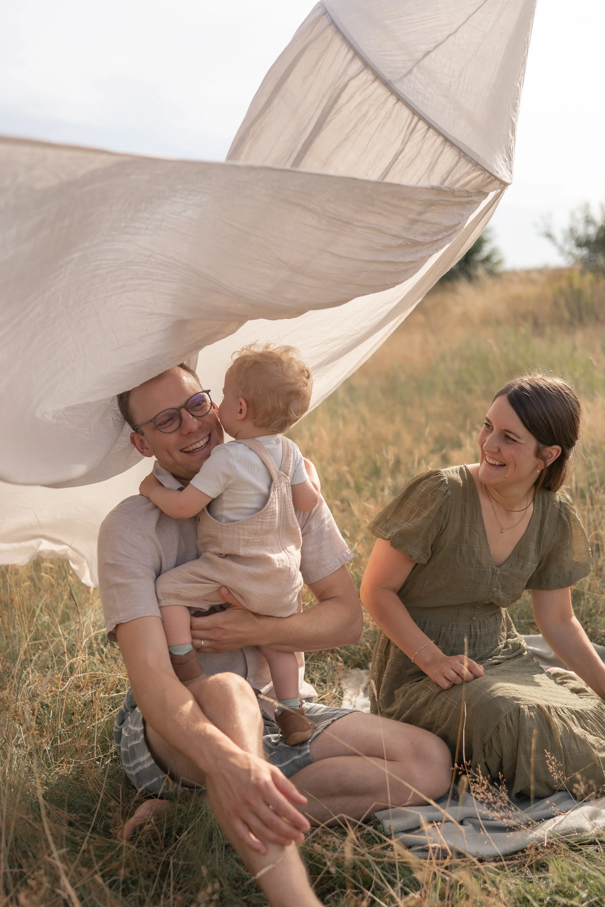 Familie im Freien auf einer Wiese, lachend und umgeben von trockenem Gras, unter einem Tuch oder einer Decke, bei schönem Wetter.