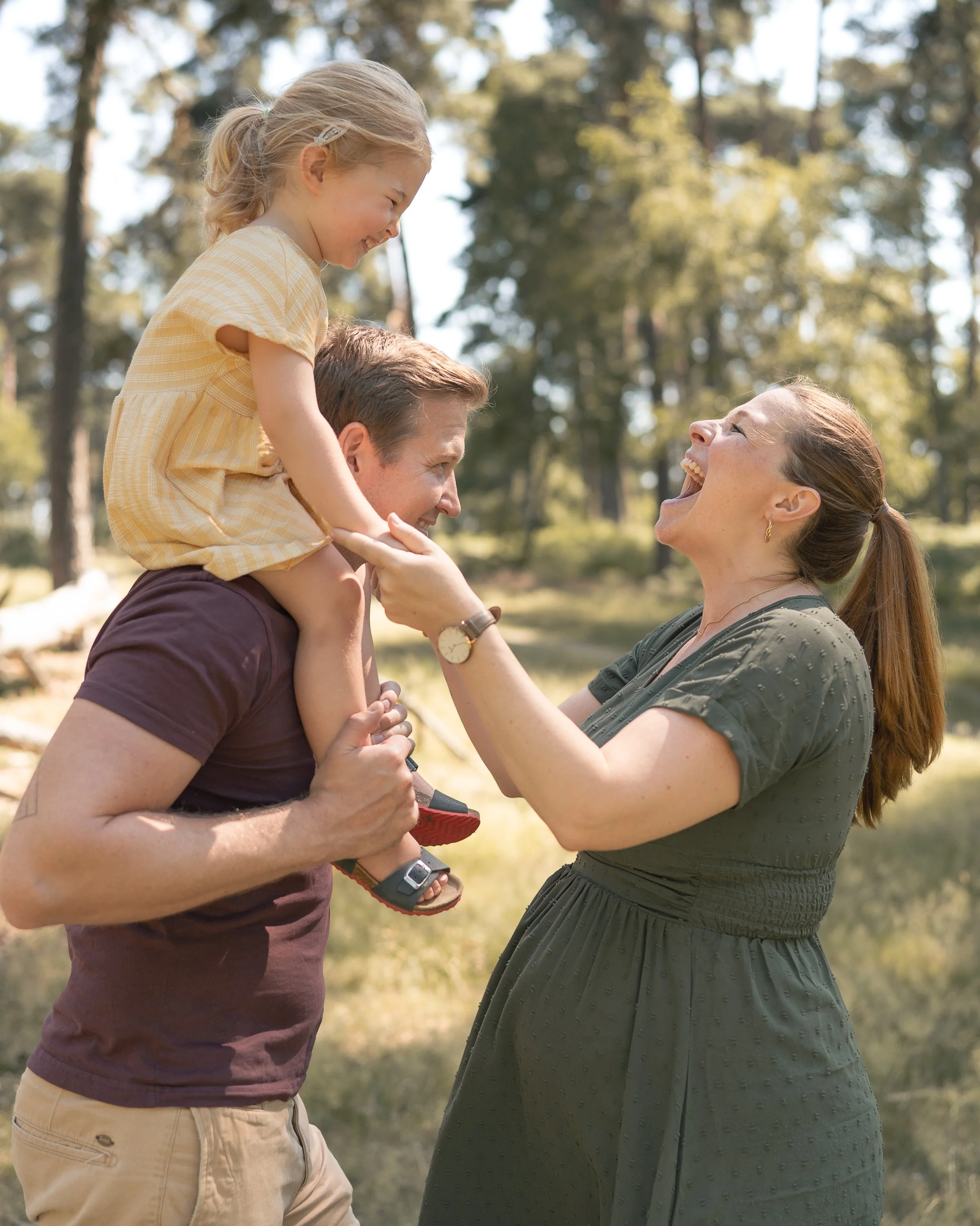 Ein Baby wird von einem Mann auf den Schultern gehalten. Eine Frau steht vor ihm und alle lachen und haben Spaß im Freien, umgeben von Bäumen bei schönem Wetter.