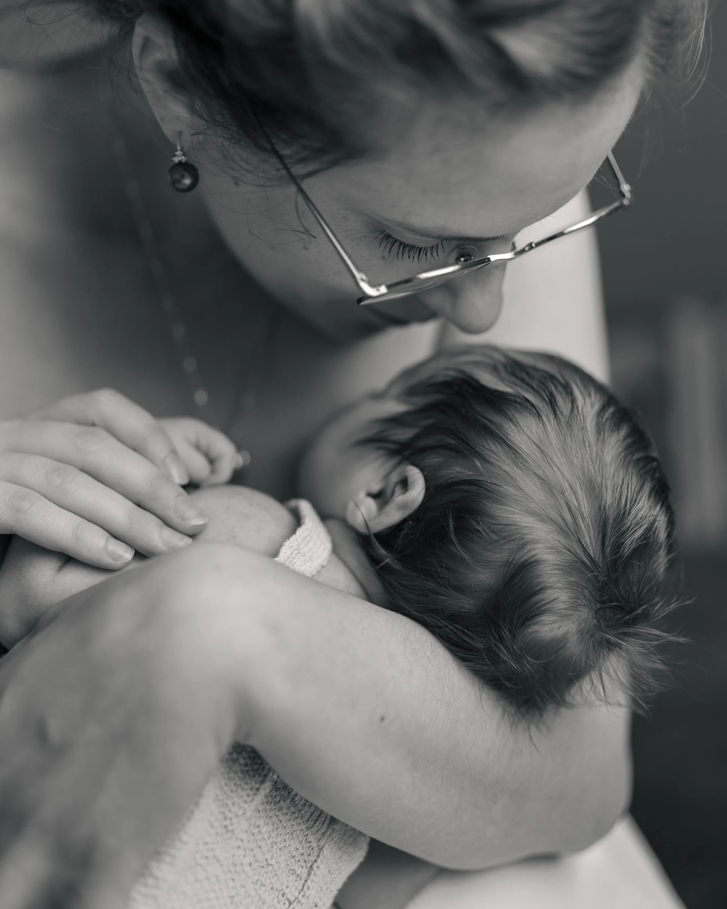 Nahaufnahme eines Babys, das auf der Brust einer Person liegt. Eine Frau, vermutlich die Mutter, trägt eine Brille und Ohrringe, während sie das Baby hält.