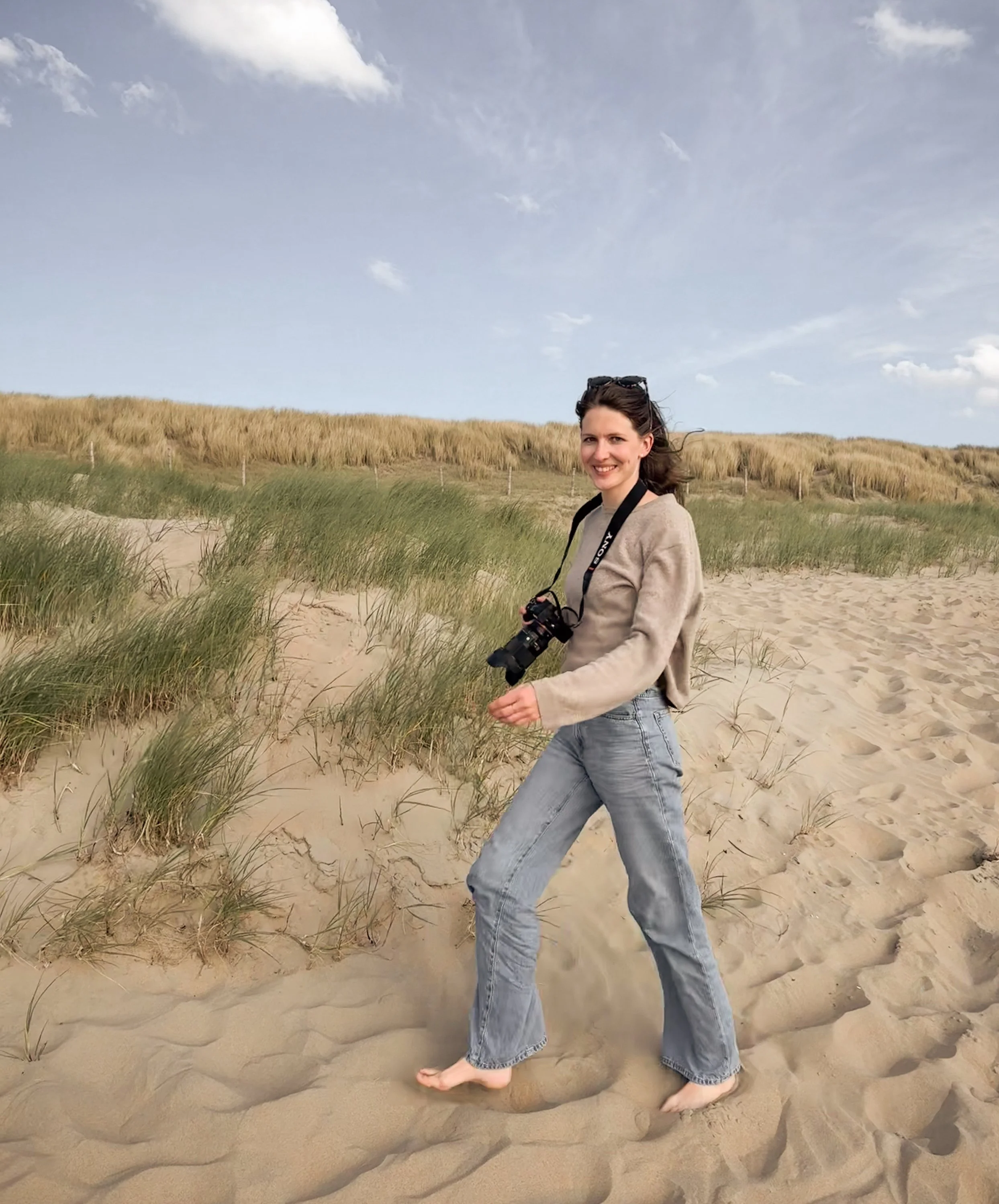 Junge Frau am Strand, barfuß im Sand, mit Kamera um den Hals, blickt lachend in die Kamera, im Hintergrund Dünen und Himmel mit Wolken.