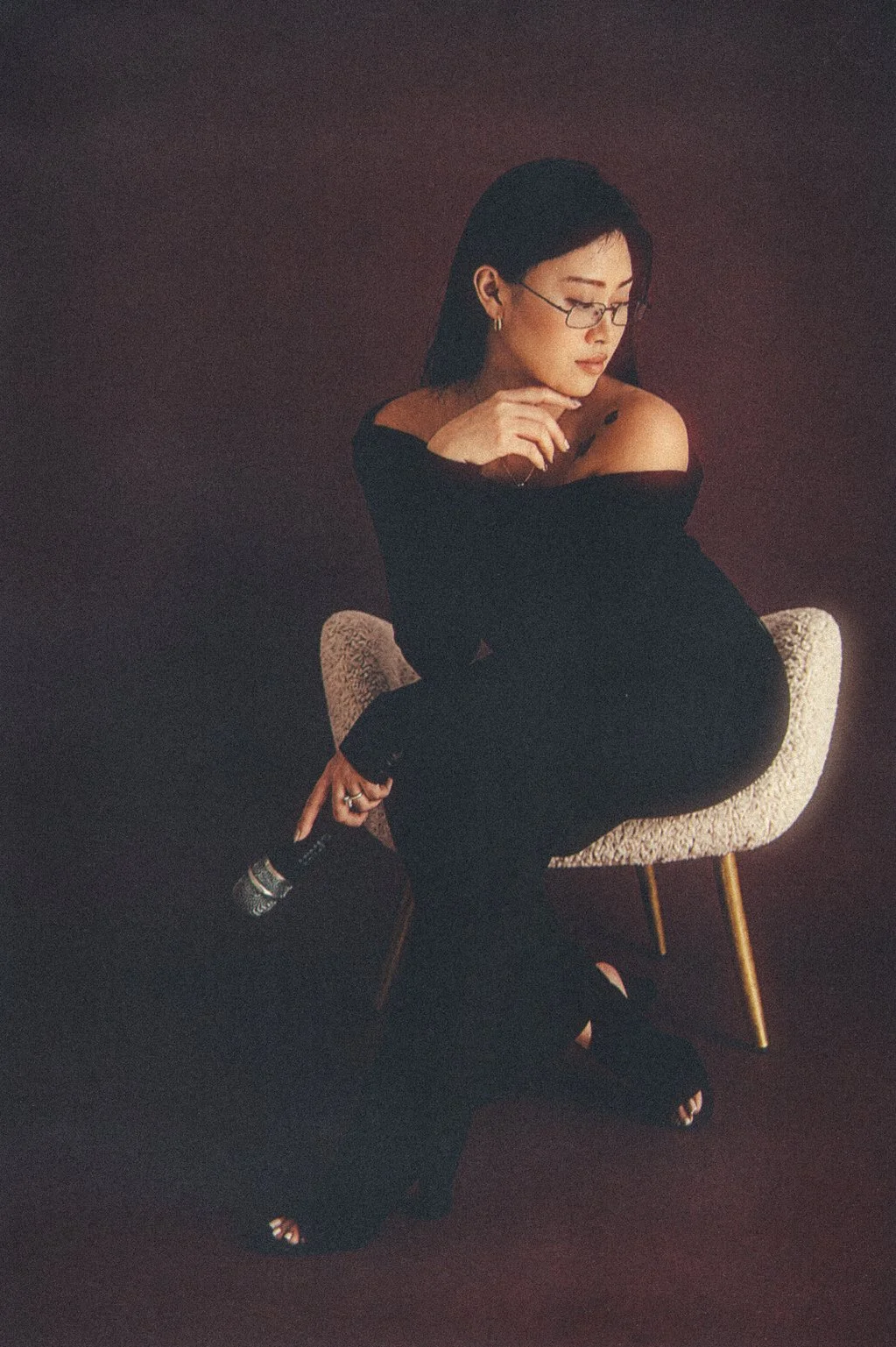 A woman with dark hair and glasses sitting on a beige chair with a maroon background. She is wearing a black off-shoulder dress and holding a microphone.