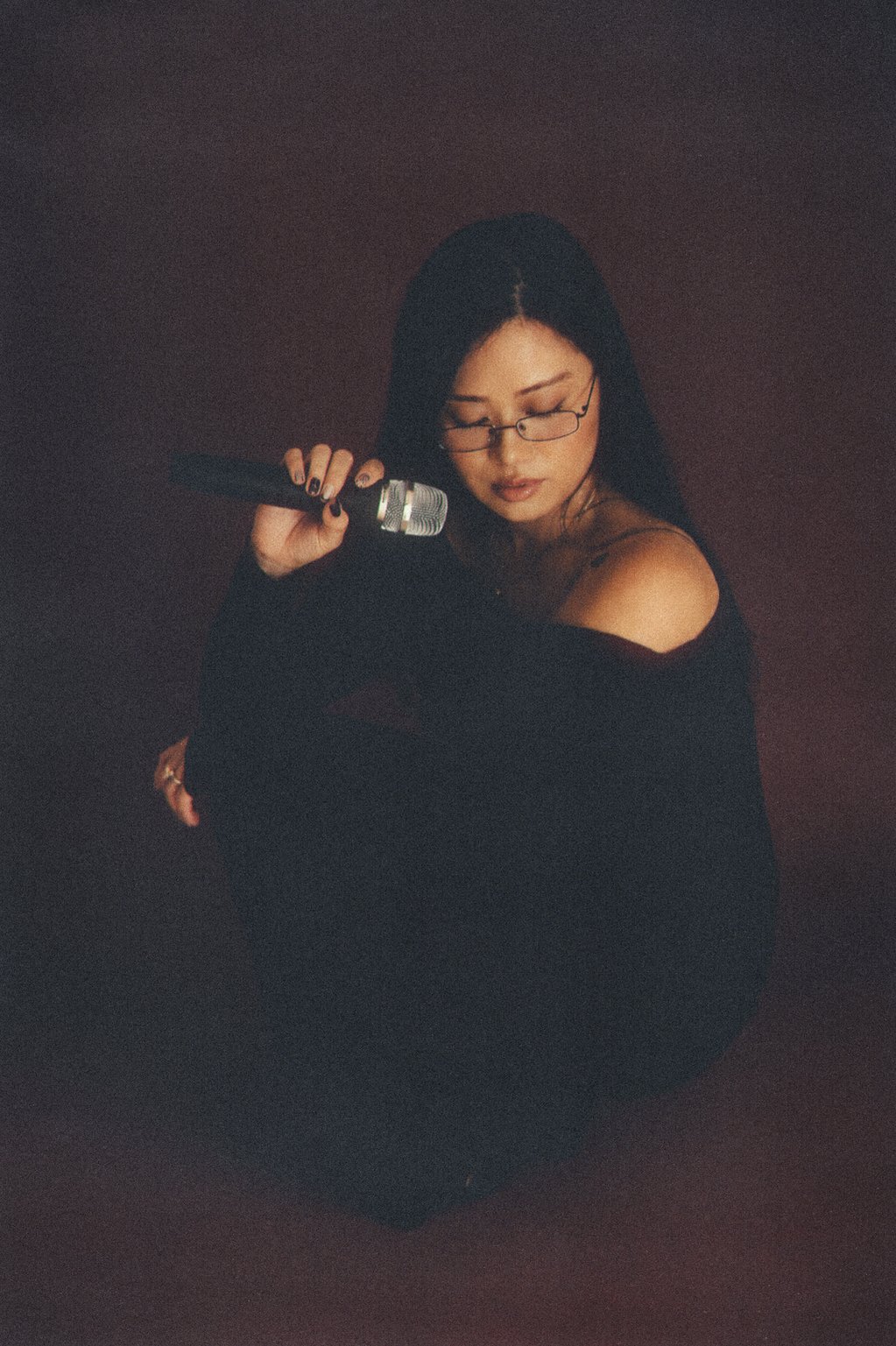 A woman with long black hair holding a microphone, wearing glasses and a black off-shoulder top, sitting against a dark background.