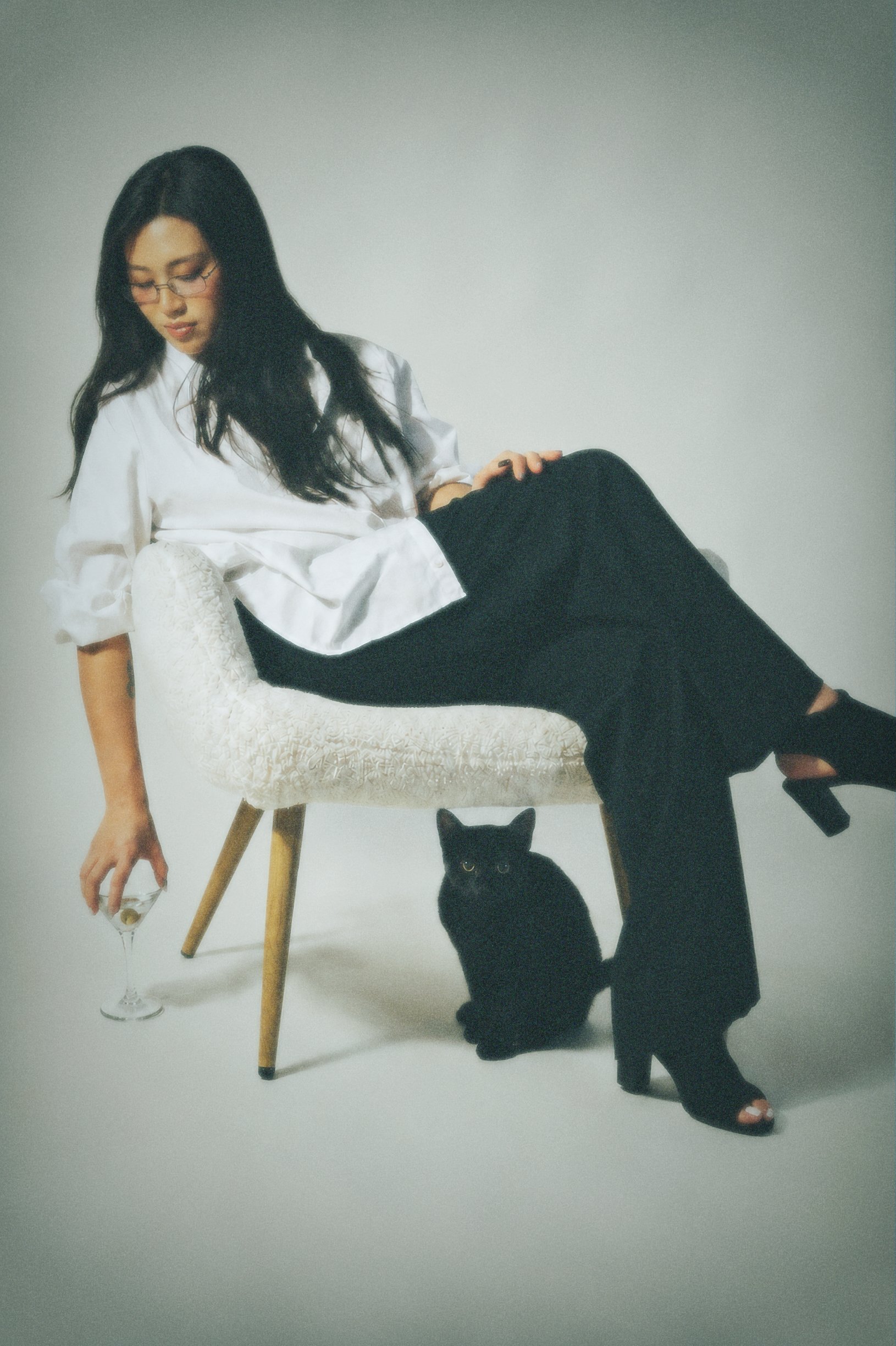 A woman with long black hair and glasses is sitting on a white cushioned stool. She is dressed in a white blouse and black pants. She is holding a glass of white wine and has a black cat sitting underneath her, looking up at her.