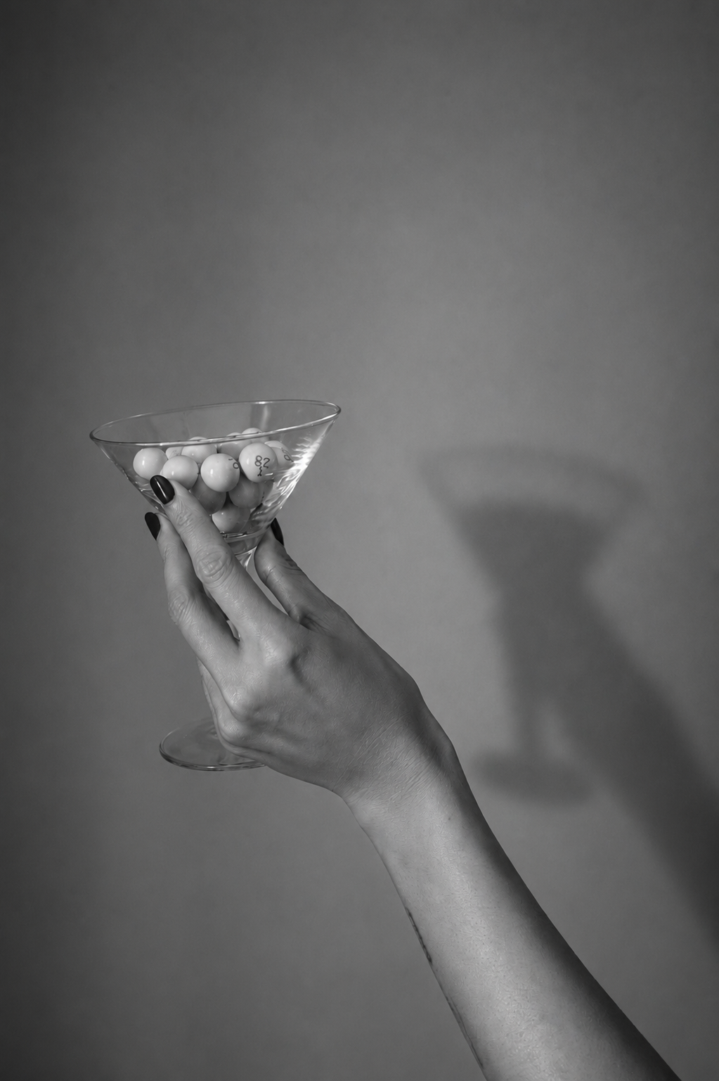 A person's hand holding a martini glass filled with bingo balls, with a plain gray background.