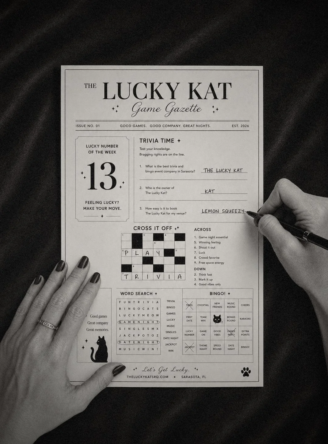 Black and white image of a person writing on a game-themed newspaper titled 'The Lucky Kat Game Gazette,' with a crossword puzzle, word search, and trivia questions, held over a black fabric background.