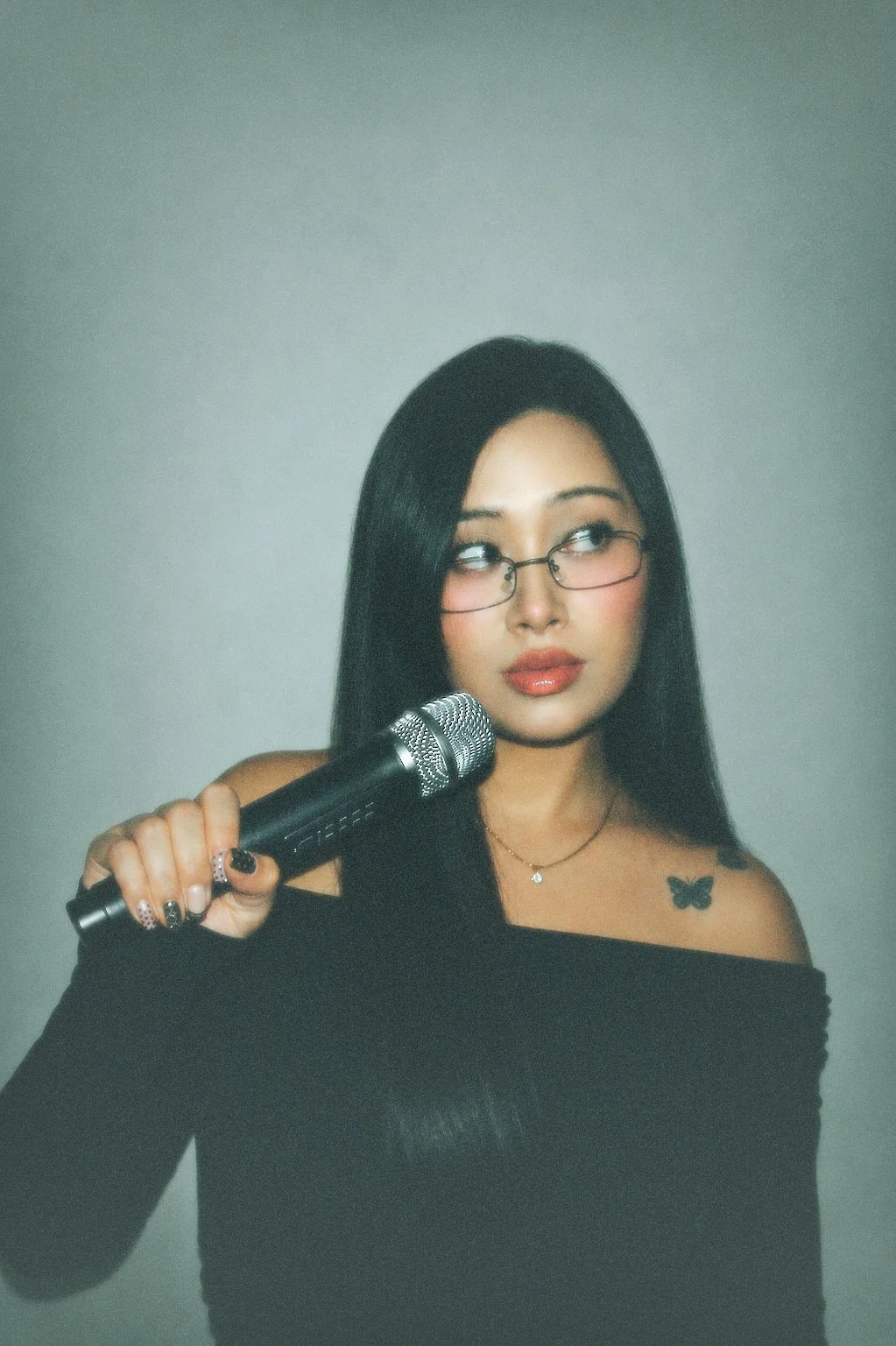 A young woman with long black hair, wearing glasses and a black off-shoulder top, holds a microphone and looks to the side with a skeptical expression. She has a butterfly tattoo on her shoulder and is wearing red lipstick and makeup.