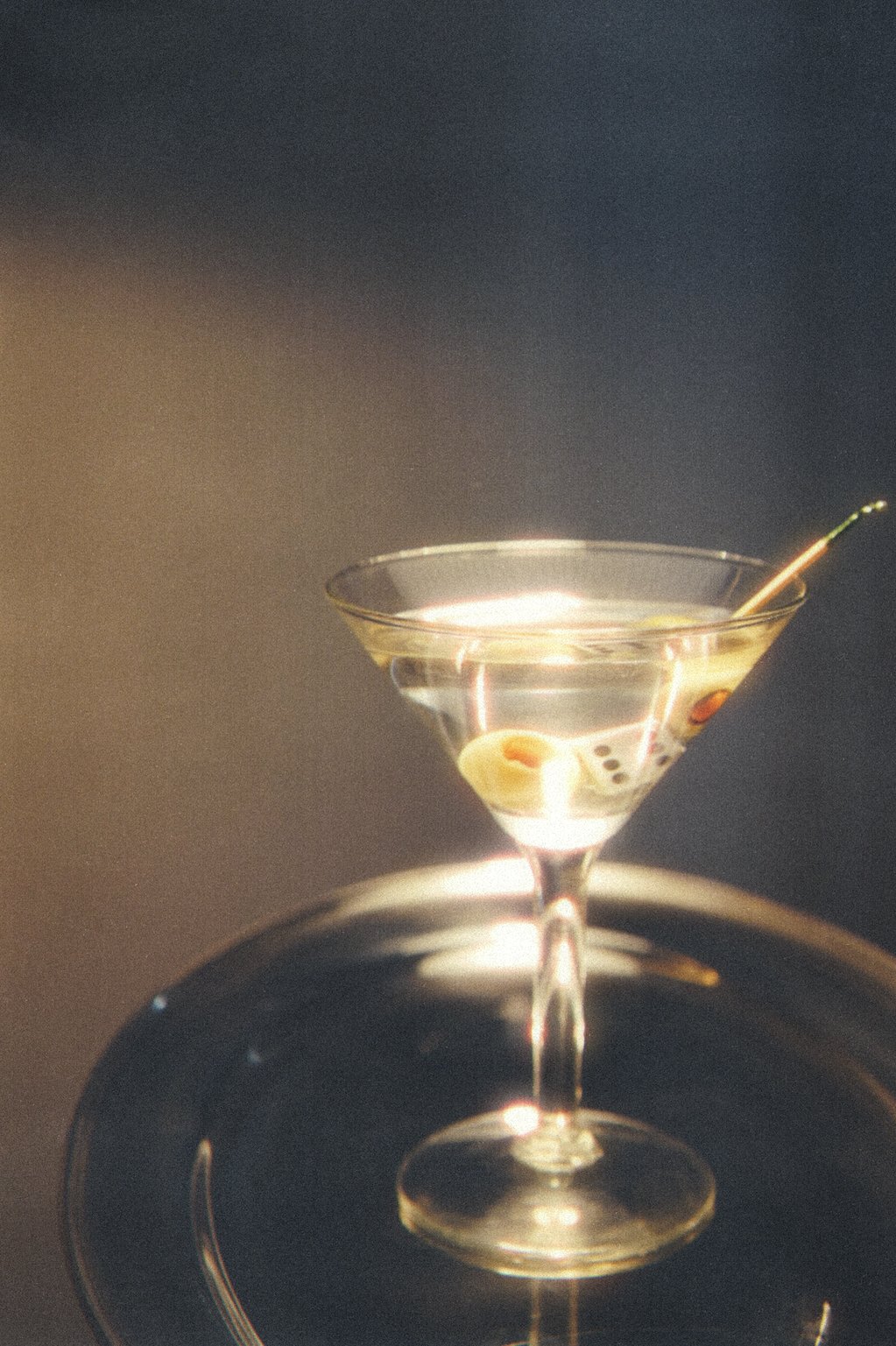 A martini glass filled with a clear cocktail, garnished with green olives on a cocktail pick, against a dark background.
