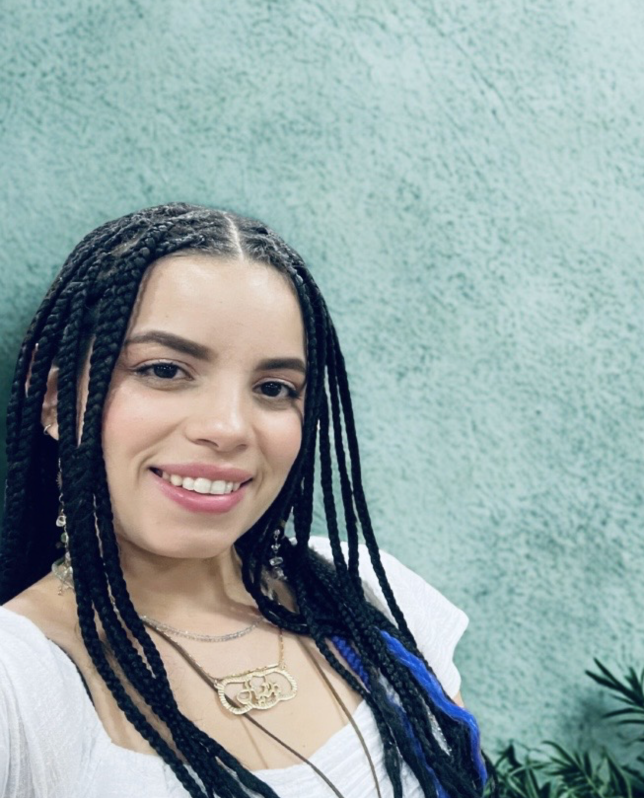 Close-up of young woman with braided hair smiling, standing against a green textured wall, wearing a white top and layered necklaces.